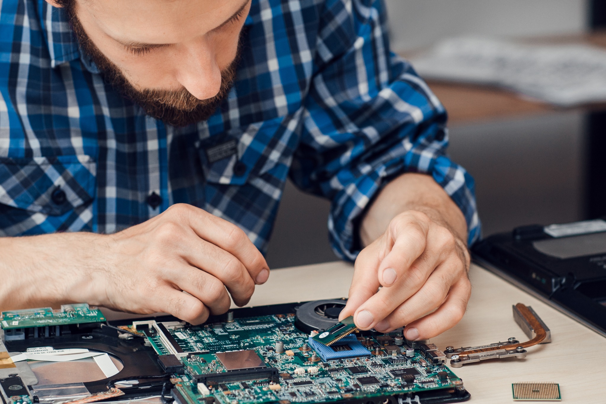 Mężczyzna w niebieskiej koszuli w kratę instaluje moduł pamięci RAM w płycie głównej laptopa, widoczne elementy chłodzenia i inne podzespoły.
