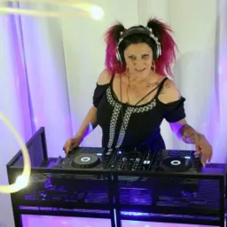 Kobieta DJ z różowymi włosami w kucykach, w słuchawkach, obsługująca sprzęt DJ-ski na tle białych zasłon z fioletowym oświetleniem.