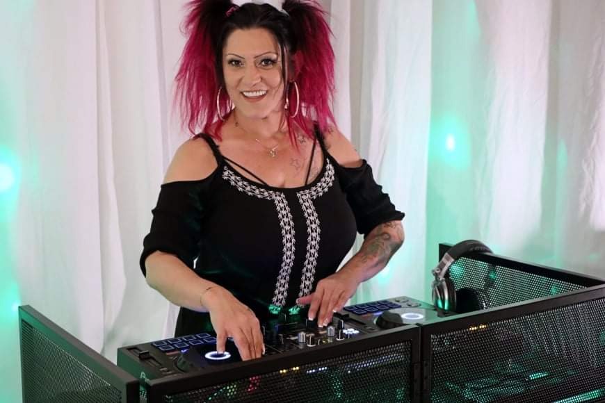 Uśmiechnięta DJ-ka z różowymi włosami w kucykach, w czarnej bluzce z odkrytymi ramionami, gra na nowoczesnym, podświetlanym kontrolerze DJ-skim na tle białych zasłon z zielonymi akcentami świetlnymi.