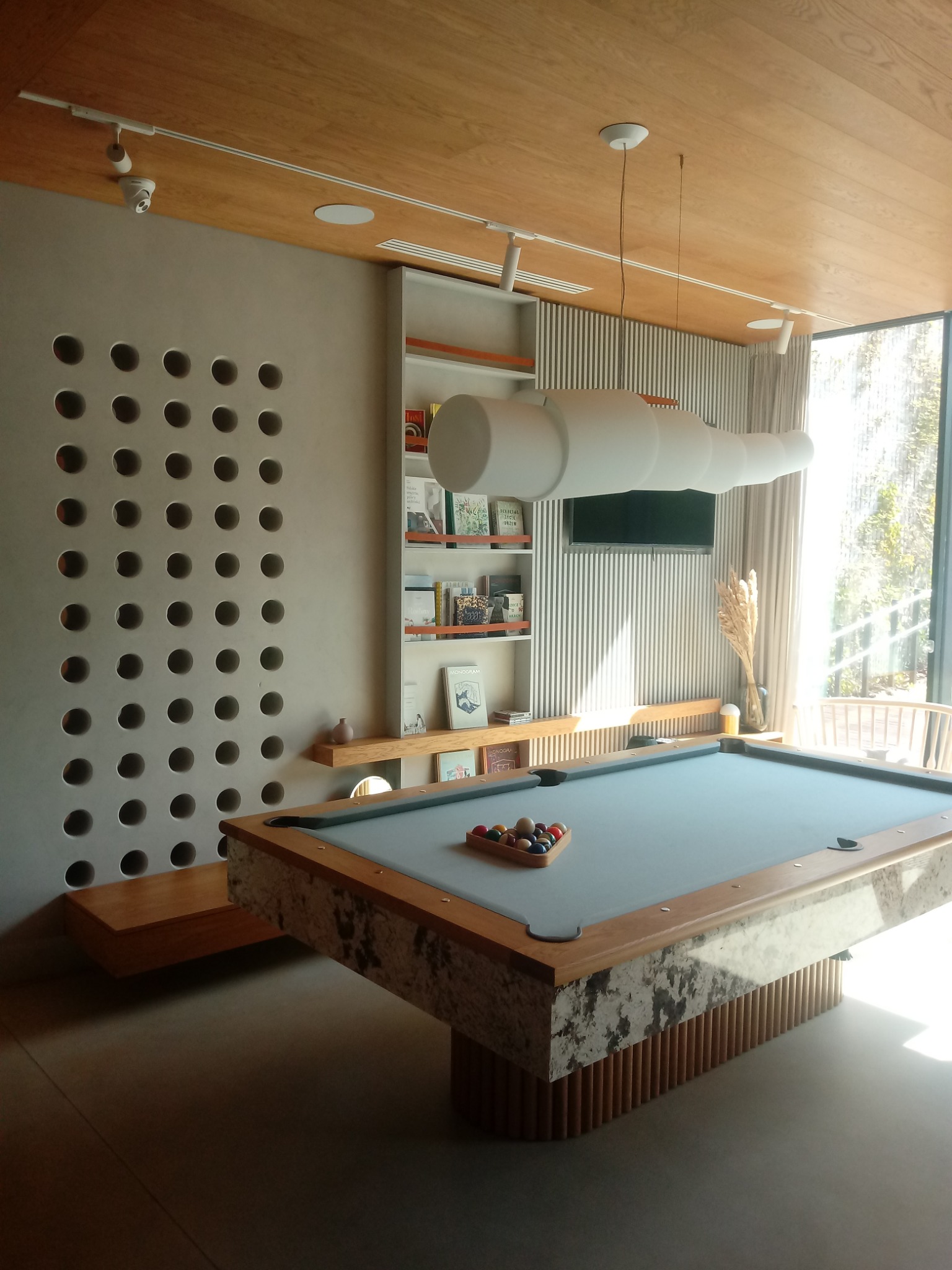 Nowoczesny pokój rekreacyjny z marmurowym stołem bilardowym, oświetleniem w kształcie tuby i ścianą z perforowanymi otworami, widok na zewnątrz przez duże okno.