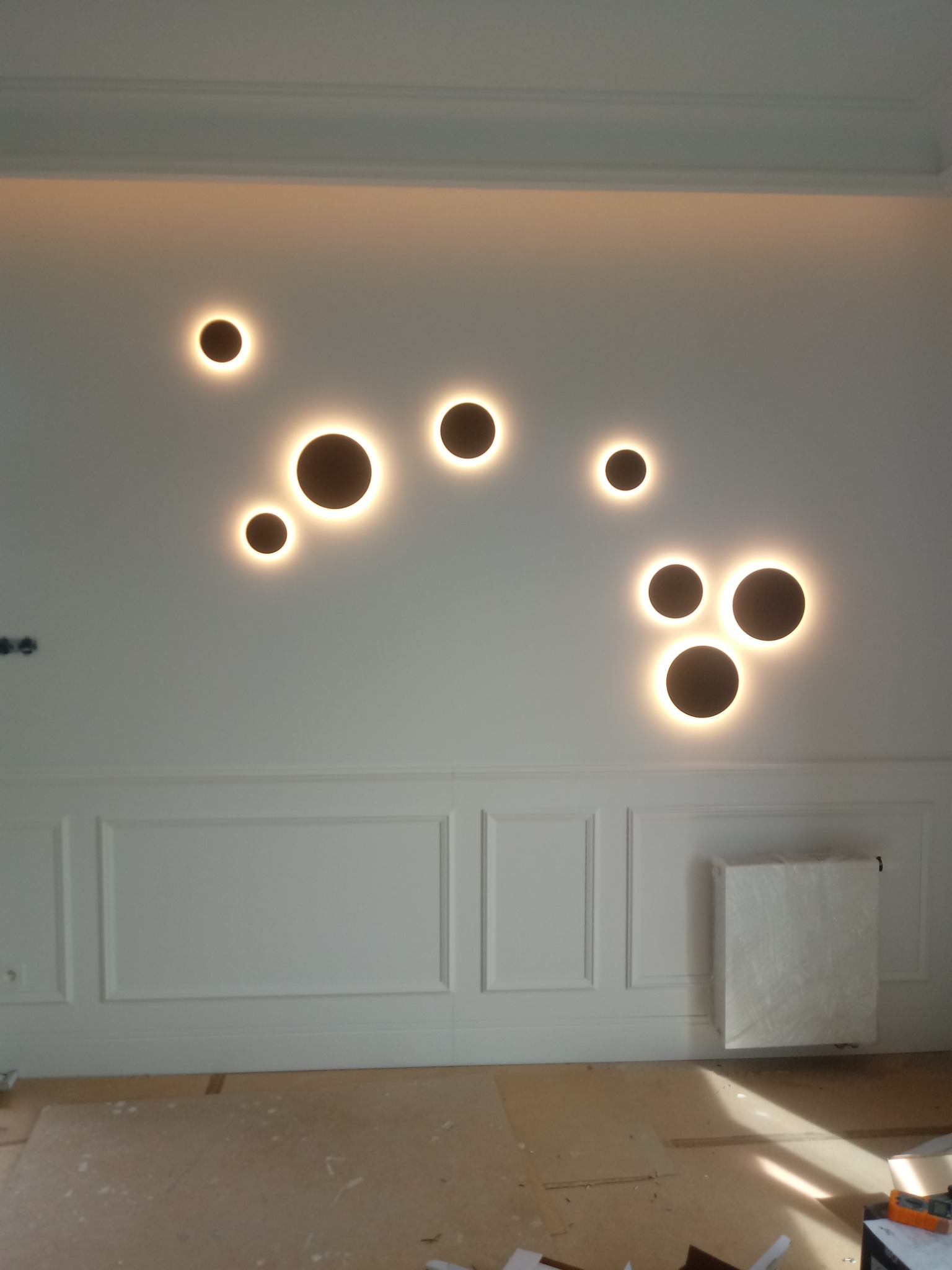 Biała ściana z dekoracyjnymi, okrągłymi lampami LED o ciepłym świetle, poniżej ozdobne panele ścienne i przykryty folią grzejnik, podłoga w trakcie prac wykończeniowych.
