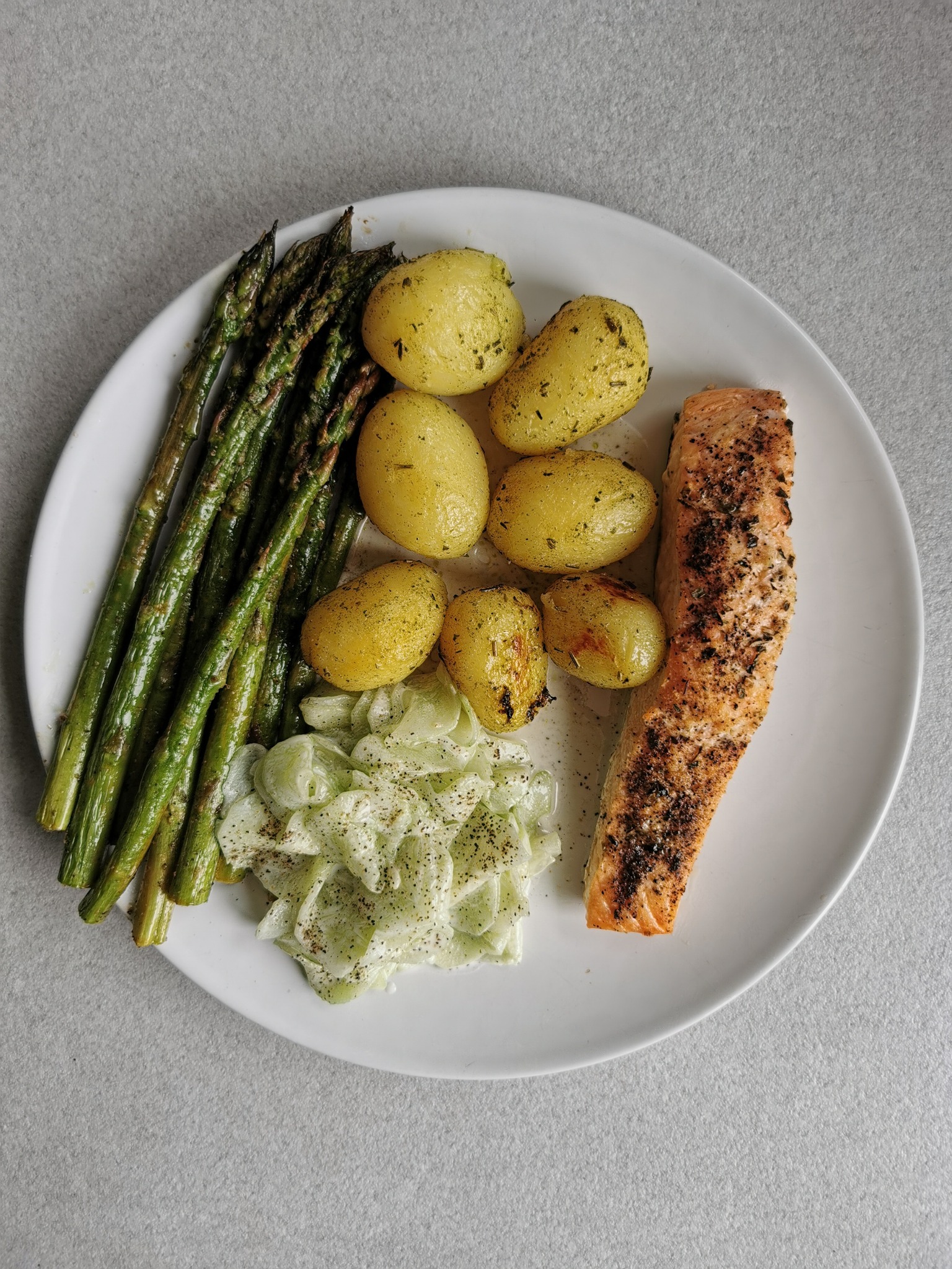 Biały talerz z grillowanym łososiem, gotowanymi ziemniakami z ziołami, szparagami i sałatką z kapusty pekińskiej z sosem na jasnoszarym tle.