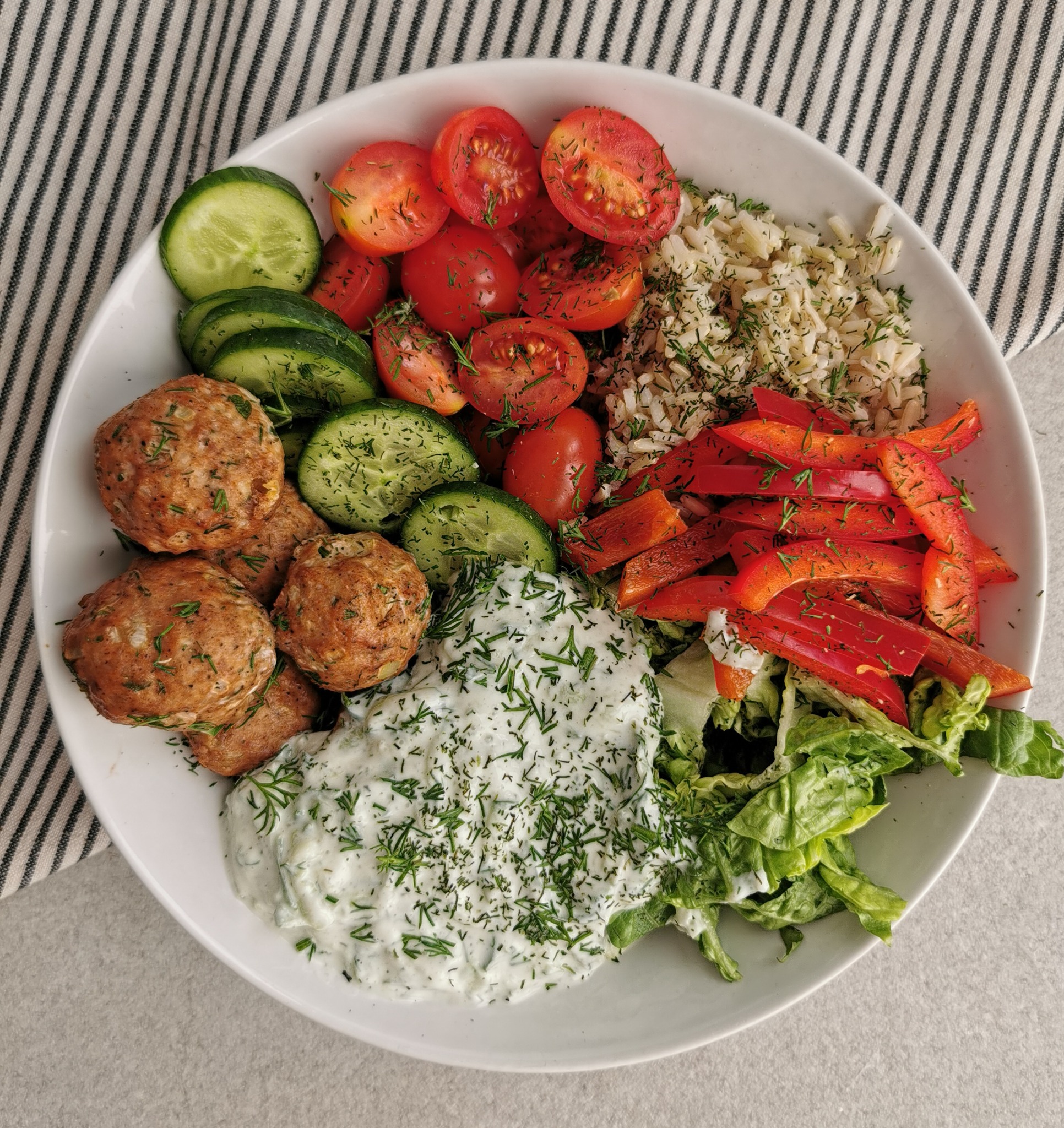 Miseczka pełna różnorodnych składników: pulpeciki, ryż, pomidorki cherry, ogórki, czerwona papryka, sałata i sos tzatziki, posypane koperkiem, na tle ściereczki w paski.