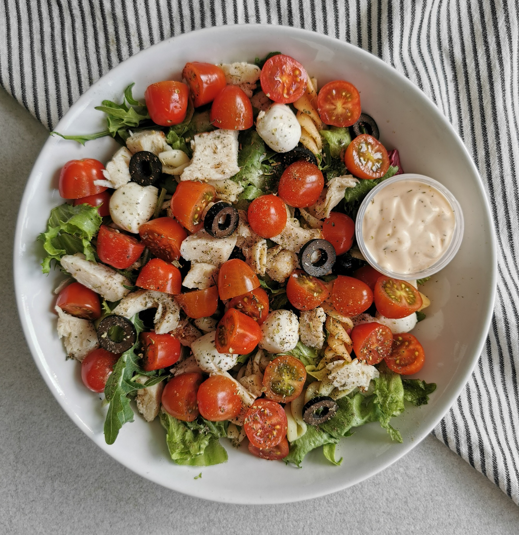 Sałatka z pomidorkami koktajlowymi, mozzarellą, oliwkami i sosem w białej misce na tle szarego blatu i ściereczki w czarno-białe paski, widok z góry.