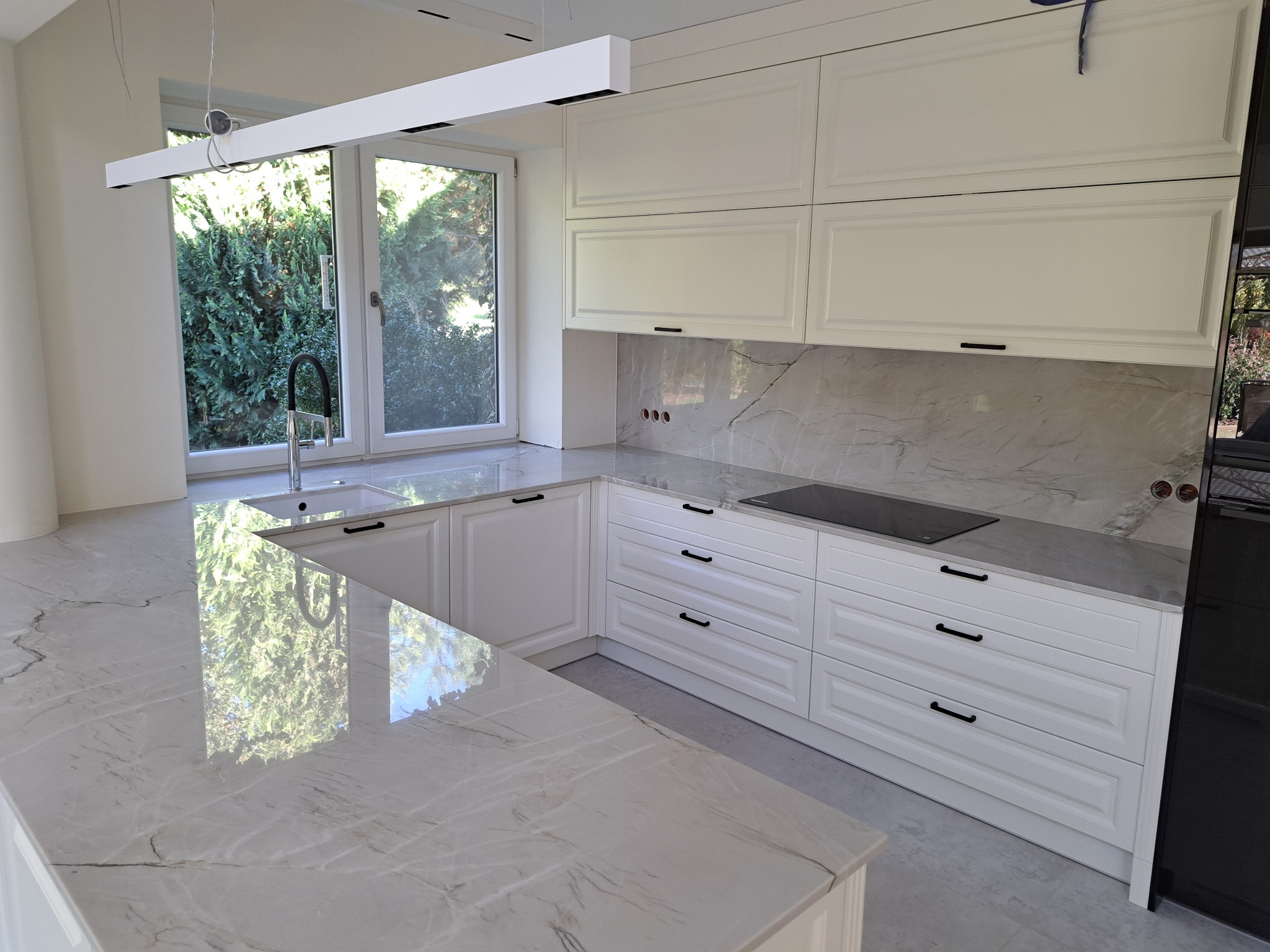 Jasna kuchnia z białymi szafkami i blatem z kamienia naturalnego z wyraźnym żyłowaniem, widok na zewnątrz przez okno nad zlewem.