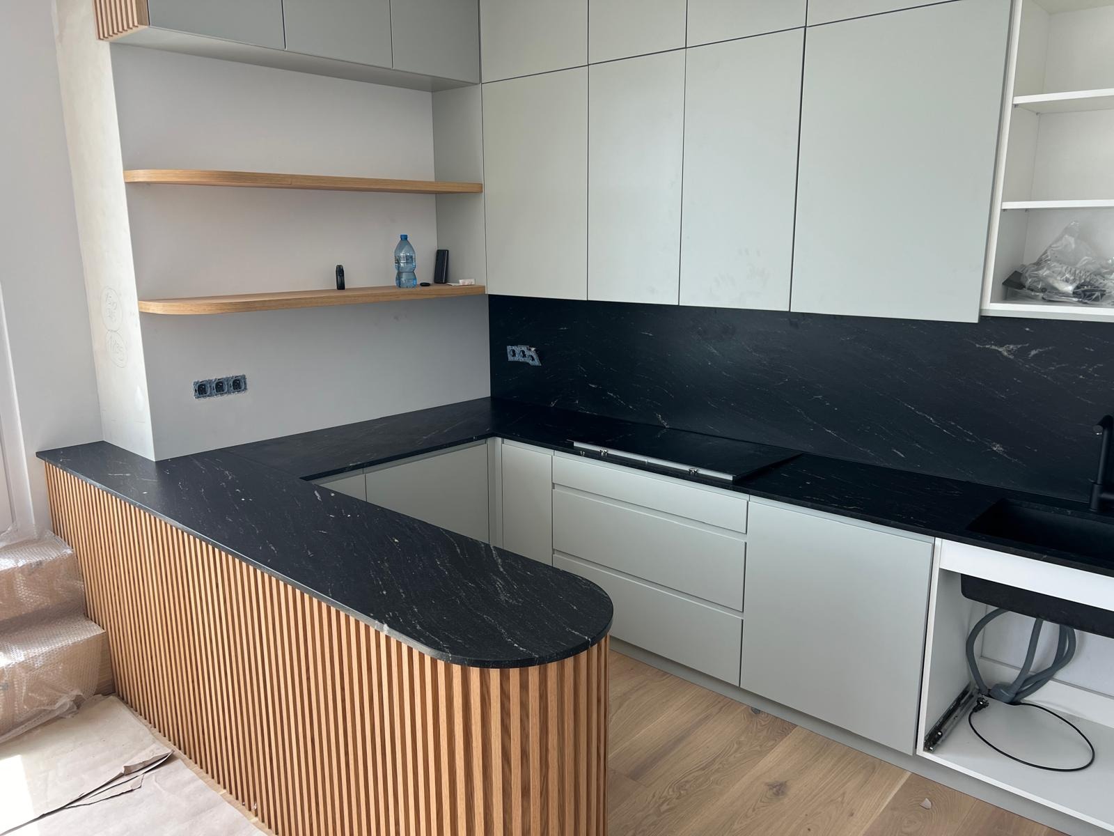 Nowoczesna kuchnia z czarnym blatem z kamienia naturalnego i drewnianym wykończeniem wyspy, jasne szafki i drewniane półki, widoczne gniazdka elektryczne.