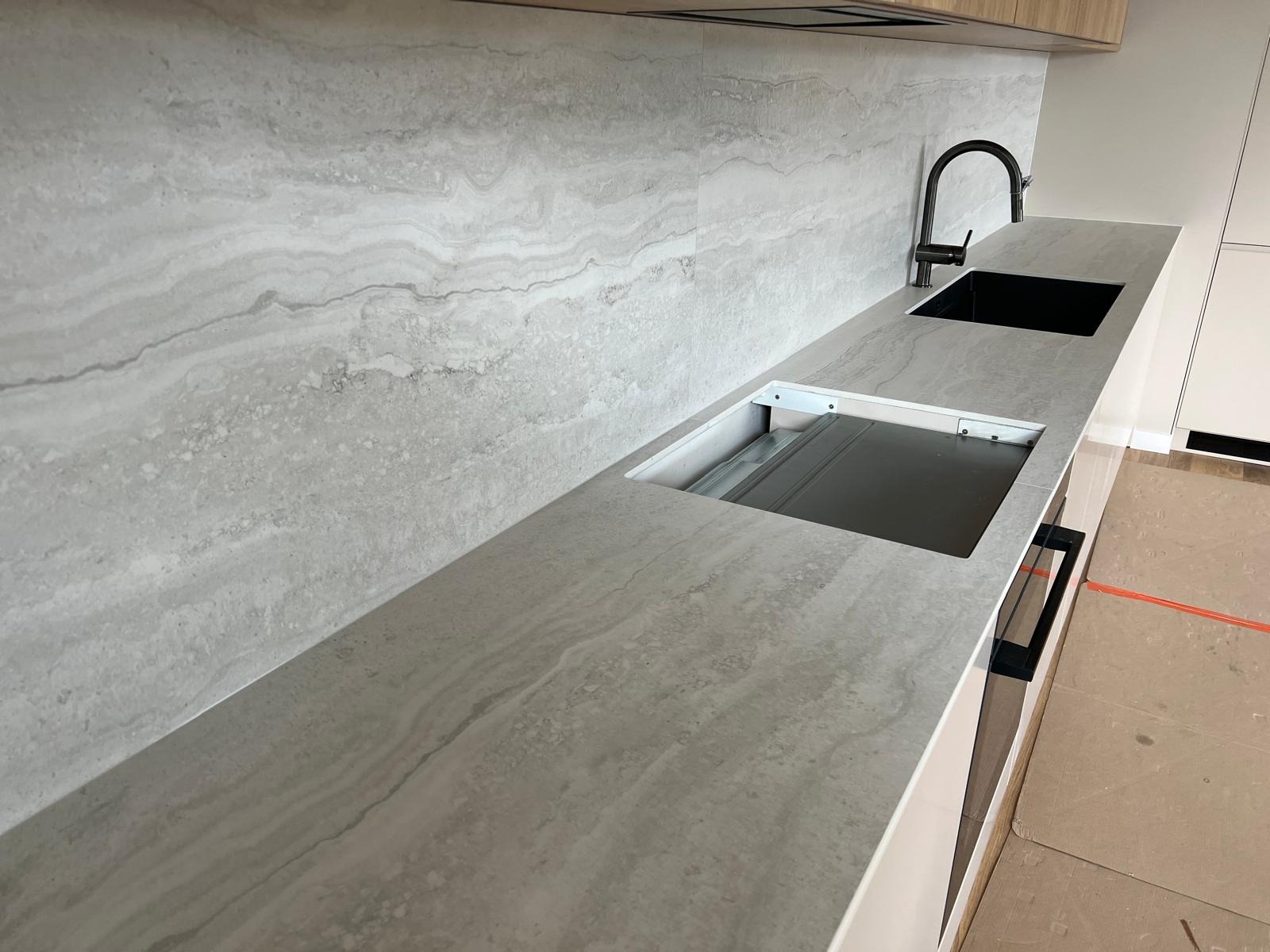 Jasnoszary blat kuchenny z wyciętym otworem na płytę grzewczą i czarnym zlewem, pasujący do ściany z płytek o podobnym wzorze.