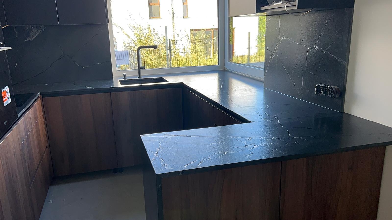 Kuchenny blat kamienny z czarnego kamienia z wyraźnym, białym żyłkowaniem, dopasowany do drewnianych szafek, z czarną armaturą i widokiem na zieleń za oknem.