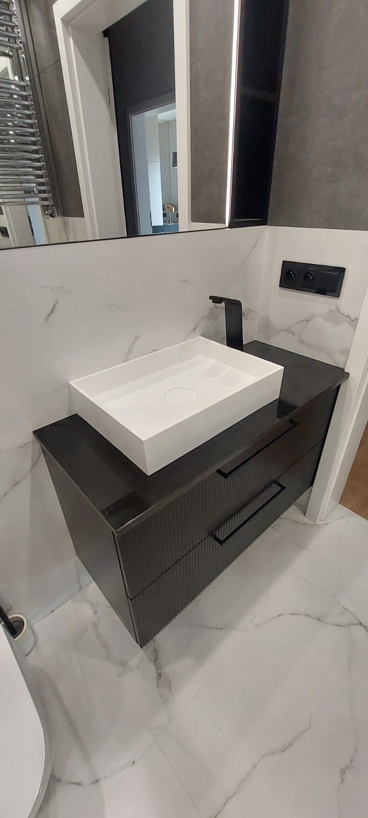 Nowoczesna łazienka z białą, prostokątną umywalką nablatową i czarną armaturą na szafce z ciemnego, ryflowanego drewna, marmurowe płytki na podłodze i ścianie.