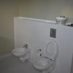 Nowoczesna łazienka z białymi, drobnymi płytkami na ścianie, wisząca toaleta z otwartą klapą, bidet i umywalka z szafką, widoczne gniazdka elektryczne i puszka z farbą na górnej półce.