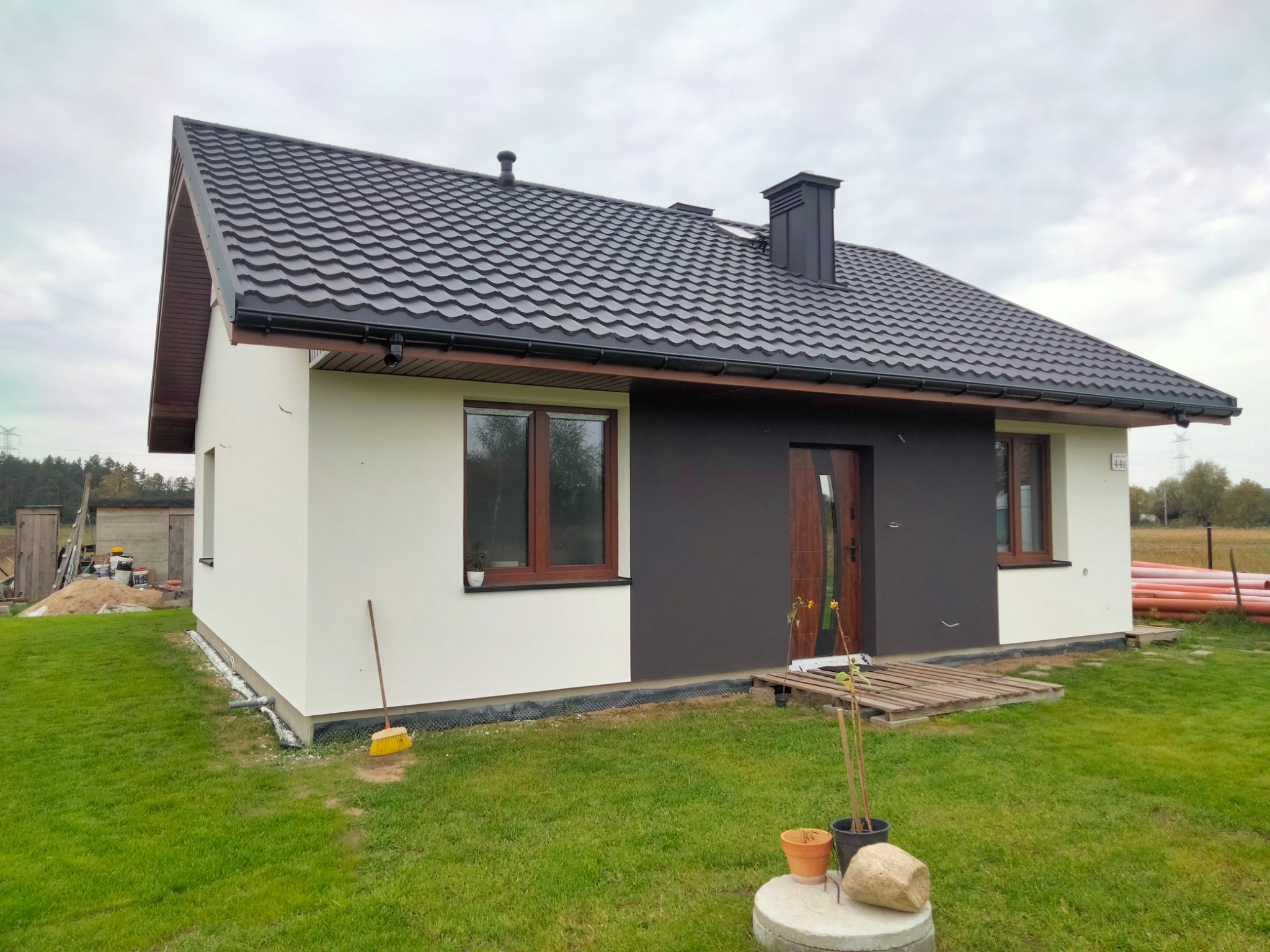 Nowoczesny dom jednorodzinny z biało-grafitową elewacją, ciemną dachówką i drewnianą stolarką okienną, widok z zewnątrz na trawnik z elementami roboczymi.