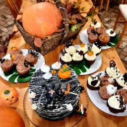 Dekoracje stołu z motywami Halloween: duża dynia w koszu, tort z pająkami i pajęczyną, muffiny z dyniami, duchami i pająkami, całość na drewnianym stole.