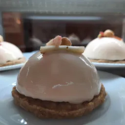 Delikatne deserki w kształcie kopuły z błyszczącą, jasnoróżową polewą na kruchym spodzie, udekorowane kawałkami kandyzowanego rabarbaru i orzechem laskowym, prezentowane na białych talerzykach...