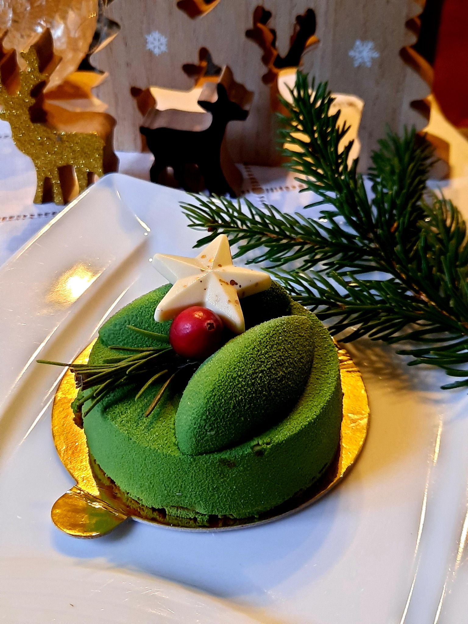 Zielone świąteczne ciastko z białą gwiazdą, czerwoną jagodą i gałązką świerku na białym talerzu, w tle drewniane renifery.