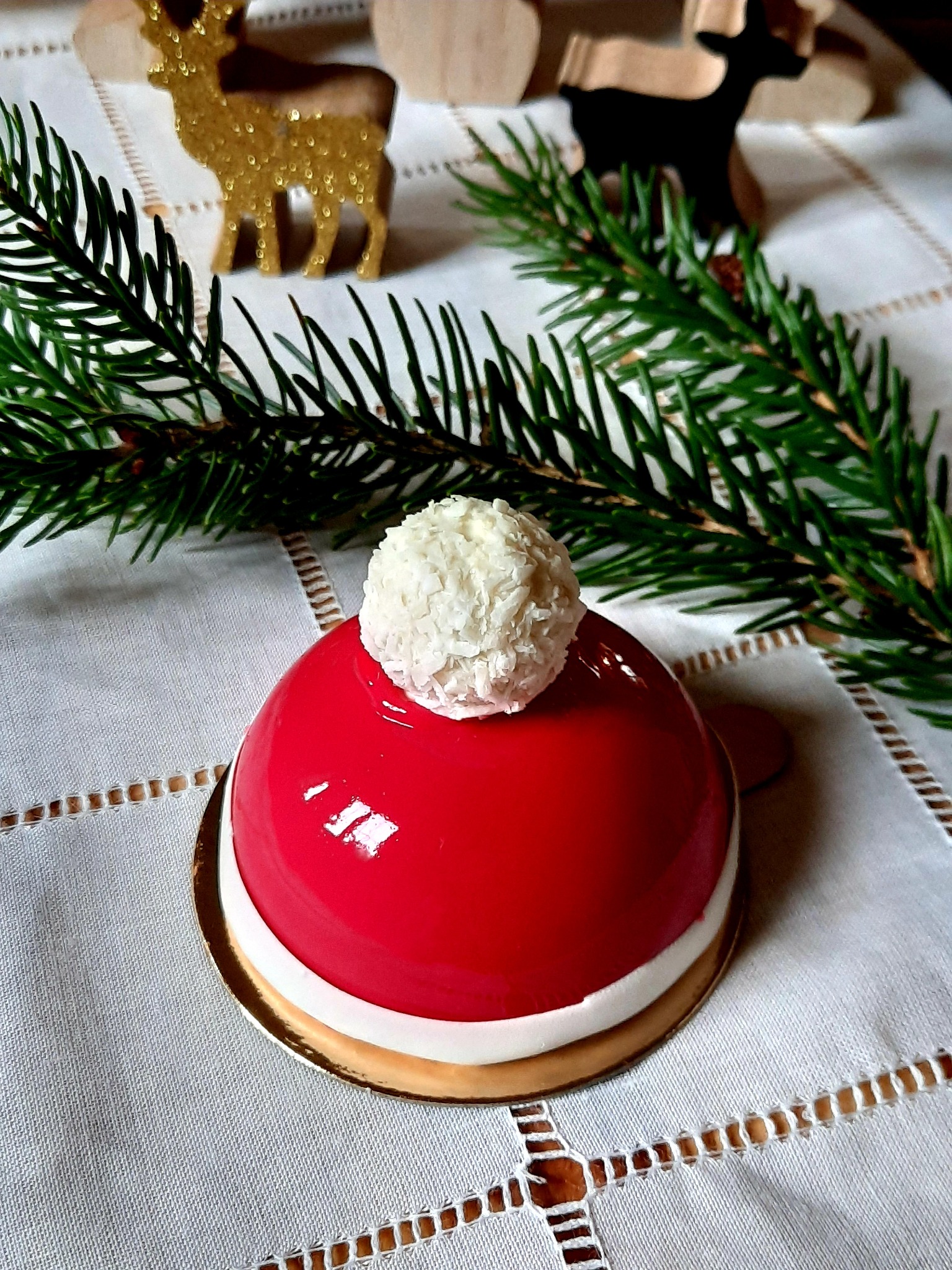 Czerwone ciastko w kształcie bombki z kokosową kulką na wierzchu, udekorowane gałązką świerku i drewnianymi ozdobami na białym obrusie.