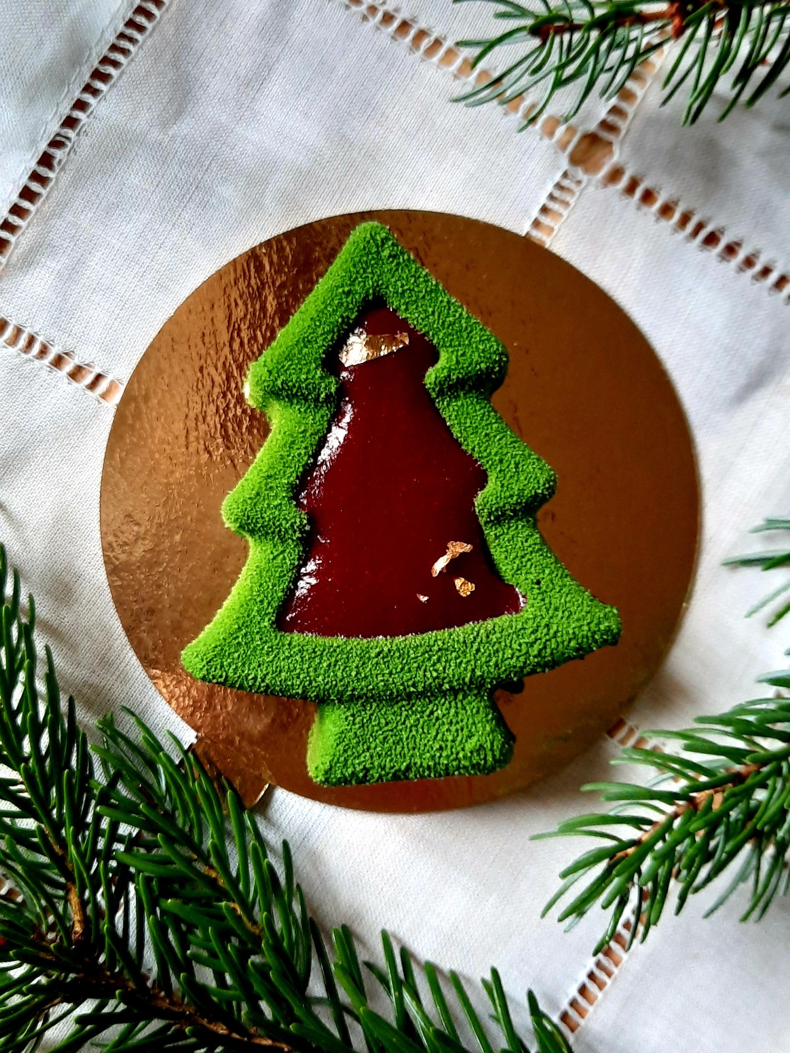 Dekoracyjny deser w kształcie choinki z zieloną, teksturowaną obwódką i czerwonym, błyszczącym wypełnieniem, ozdobiony płatkami złota, umieszczony na złotej podkładce na białym obrusie...