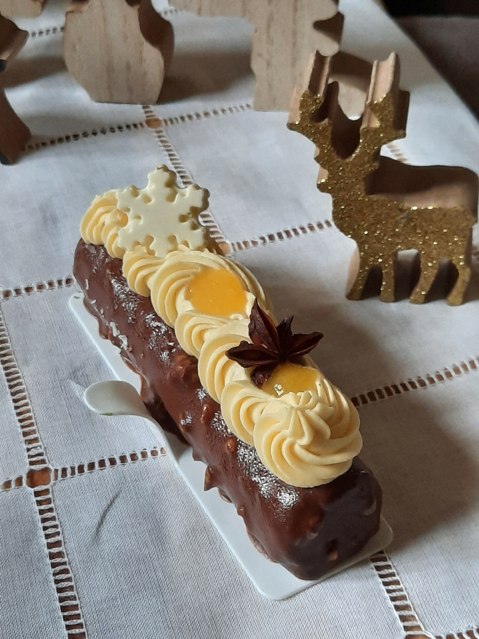 Dekoracyjny batonik czekoladowy z kremem, kandyzowaną gwiazdą anyżu i białą czekoladową śnieżynką na białym talerzyku, obok drewniany renifer z brokatem, na białym obrusie z ażurowym wzorem.