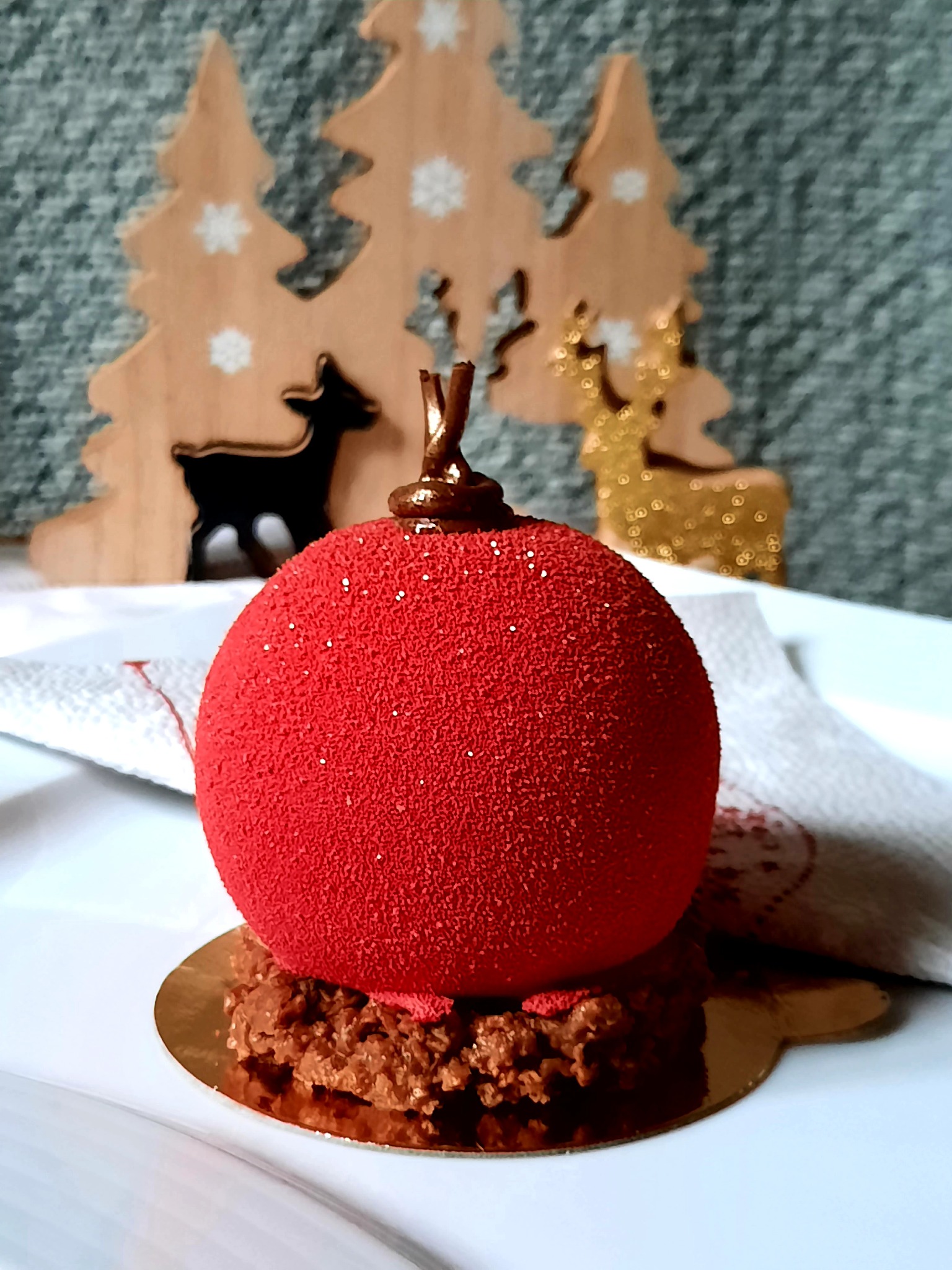 Czerwony deser w kształcie bombki z czekoladową podstawą, na białym talerzu, z dekoracjami świątecznymi w tle.