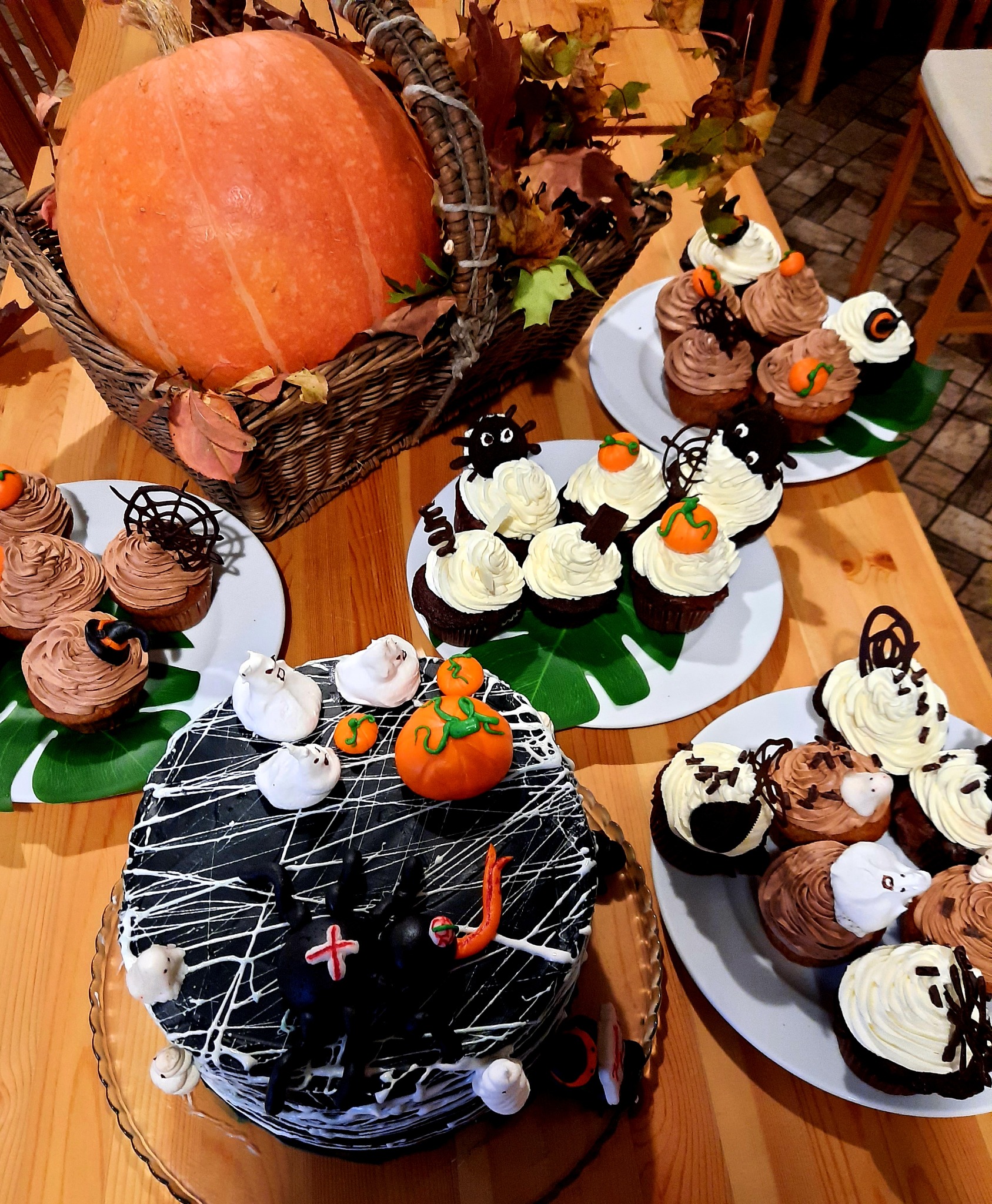 Dekoracje Halloween: duża dynia w koszyku z liśćmi, tort z pająkiem i duchami z lukru oraz babeczki z motywami dyni, pająków i duchów na drewnianym stole.