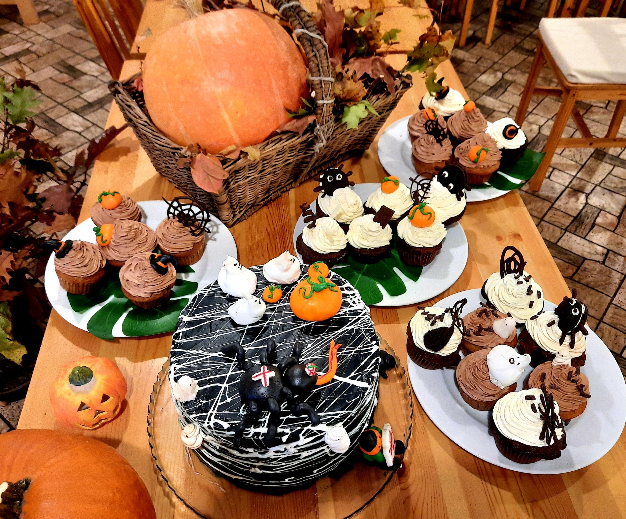 Dekoracje stołu z motywami Halloween: duża dynia w koszu, tort z pająkami i pajęczyną, muffiny z dyniami, duchami i pająkami, całość na drewnianym stole.