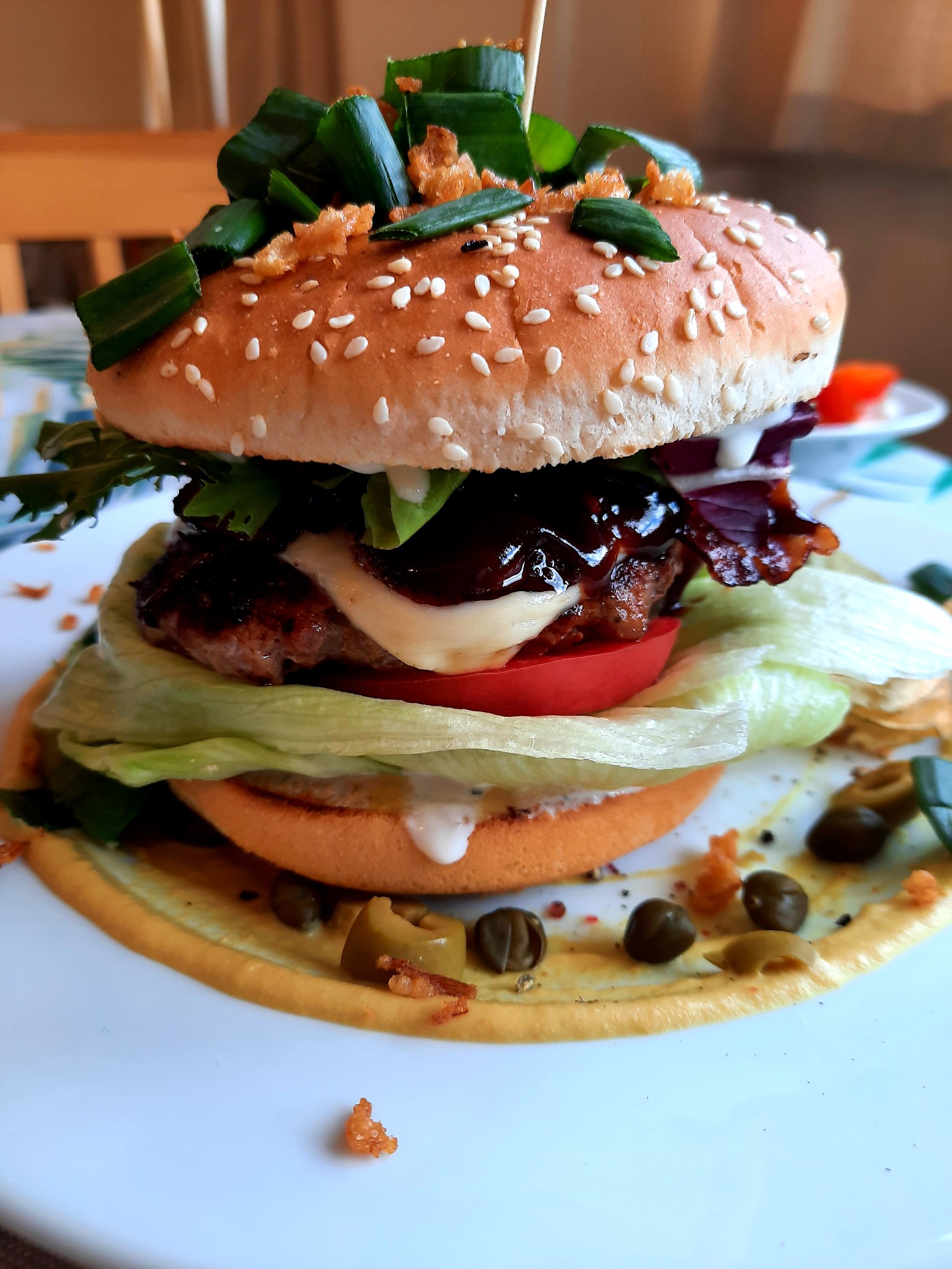 Apetyczny burger z wołowiną, serem, pomidorem, sałatą, czerwoną cebulą, sosem BBQ i szczypiorkiem na bułce posypanej sezamem, podany na białym talerzu z sosem i kaparami.