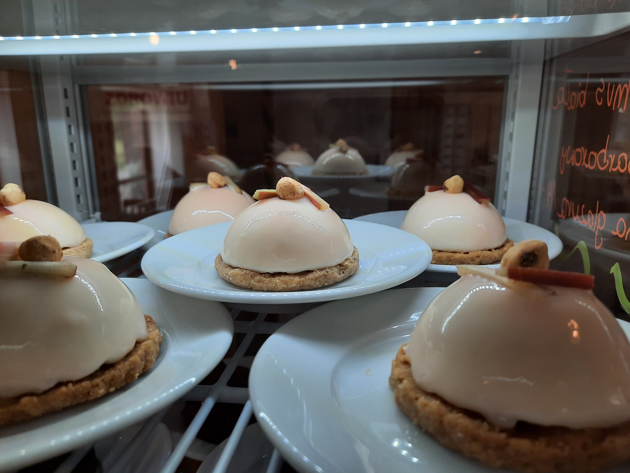 Eleganckie, kremowe desery na kruchym spodzie, udekorowane orzechem i kawałkiem kandyzowanej skórki, prezentowane w chłodniczej witrynie cukierniczej.