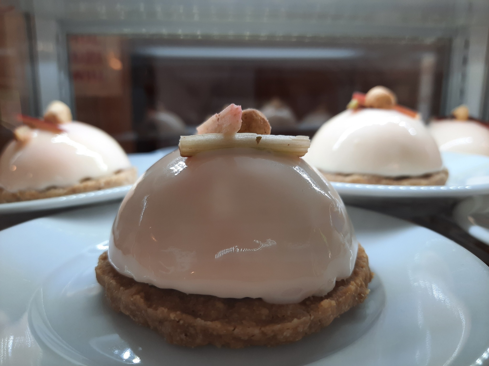 Delikatne deserki w kształcie kopuły z błyszczącą, jasnoróżową polewą na kruchym spodzie, udekorowane kawałkami kandyzowanego rabarbaru i orzechem laskowym, prezentowane na białych talerzykach...