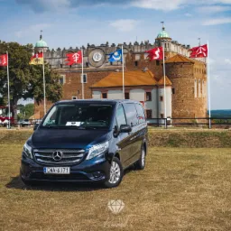 Granatowy Mercedes Vito na trawiastym placu przed zamkiem w Dobczycach, otoczonym flagami.