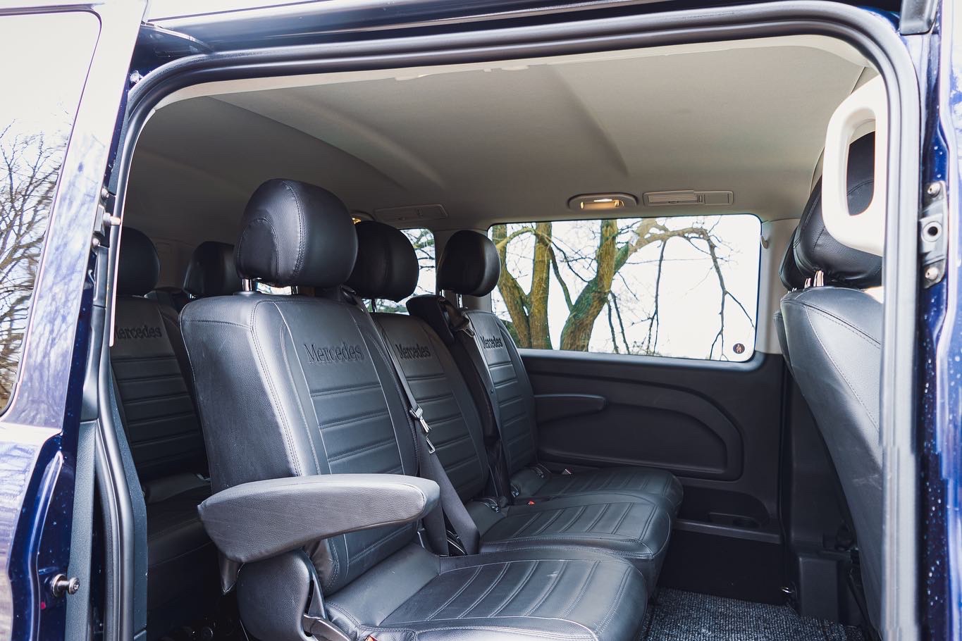 Wnętrze czarnego, skórzanego busa Mercedes z widokiem na drzewo za oknem.