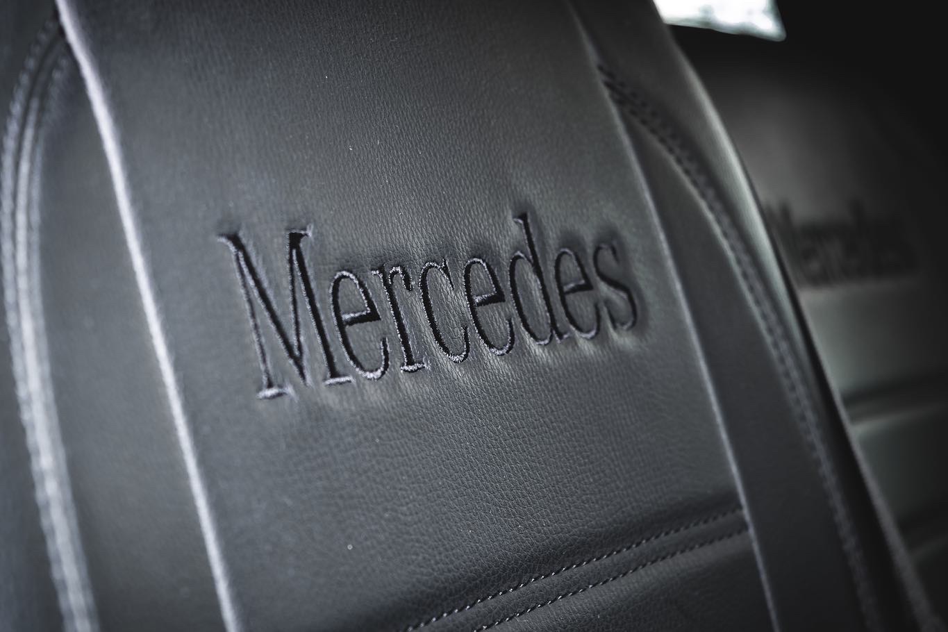 Czarne skórzane siedzenie samochodowe marki Mercedes z wyhaftowanym logo, widoczne przeszycia i pas bezpieczeństwa.