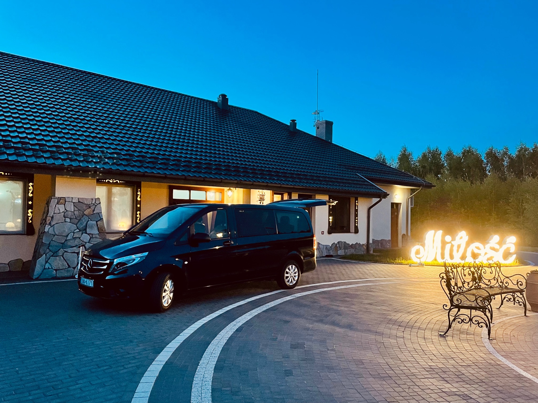 Czarny van Mercedes z otwartym bagażnikiem zaparkowany przed budynkiem z napisem 'Miłość' w dekoracyjnym oświetleniu.