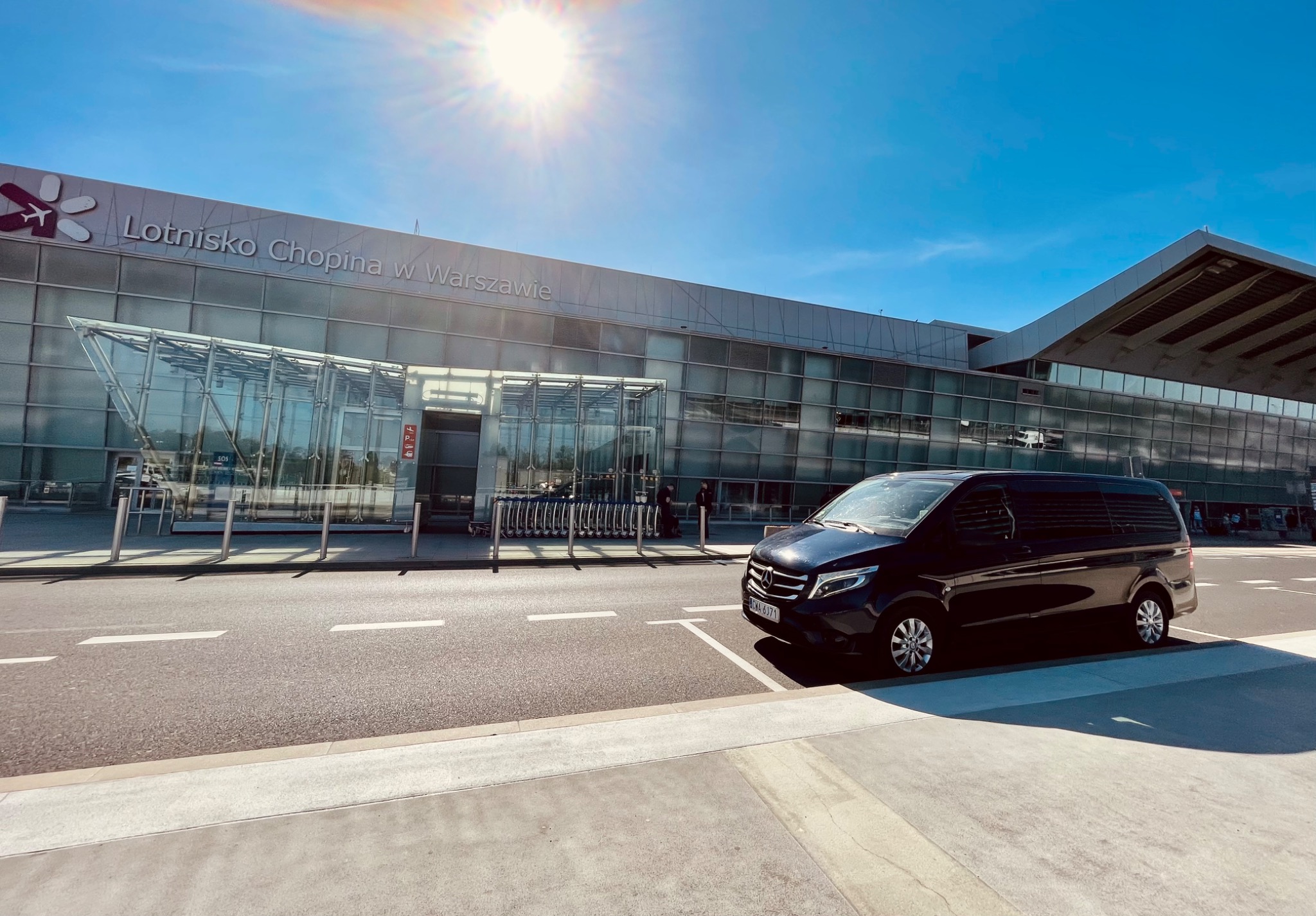 Czarny van Mercedes-Benz V-klasy zaparkowany przed lotniskiem Chopina w Warszawie, w słoneczny dzień.