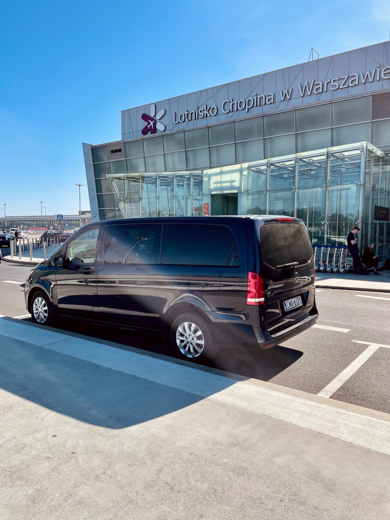 Czarny van Mercedes-Benz Vito zaparkowany przed Lotniskiem Chopina w Warszawie w słoneczny dzień.