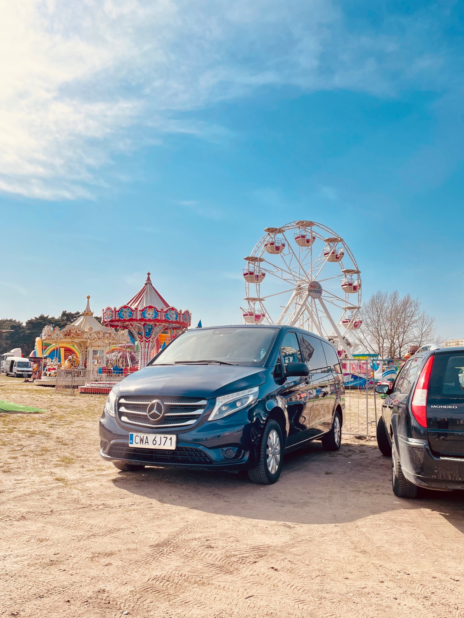 Czarny Mercedes V-klasy na piaszczystym placu zabaw z karuzelą i diabelskim młynem w tle, tablica rejestracyjna CWA 6J71.