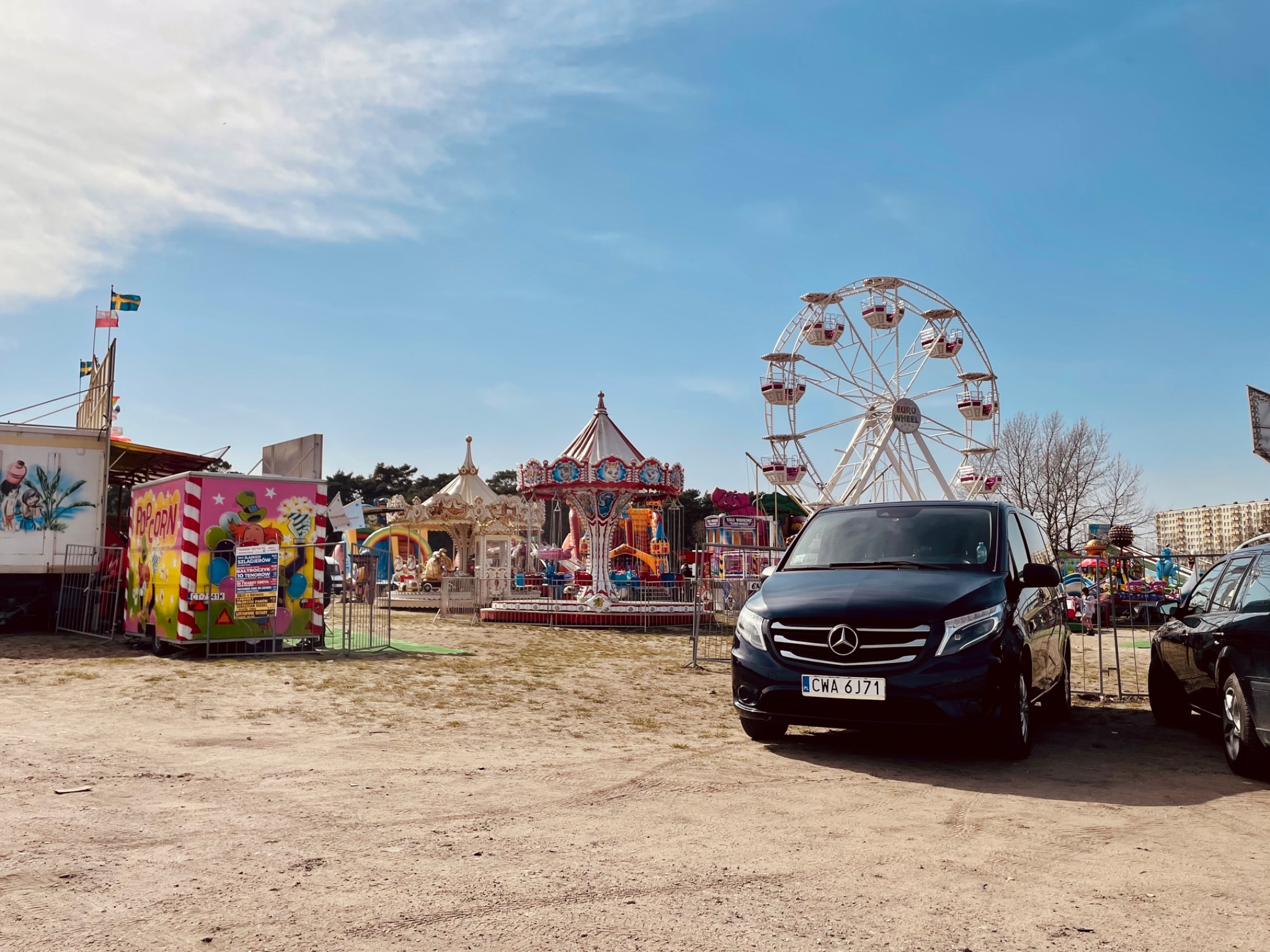 Czarny van Mercedes zaparkowany przed wesołym miasteczkiem z karuzelą i diabelskim młynem w tle.