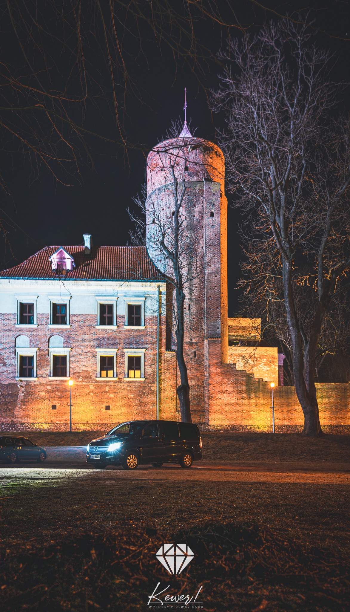 Czarny van z włączonymi światłami parkuje nocą w Wąbrzeźnie na tle oświetlonego, ceglanego zamku. Widoczny fragment drugiego samochodu i logo firmy na dole kadru.