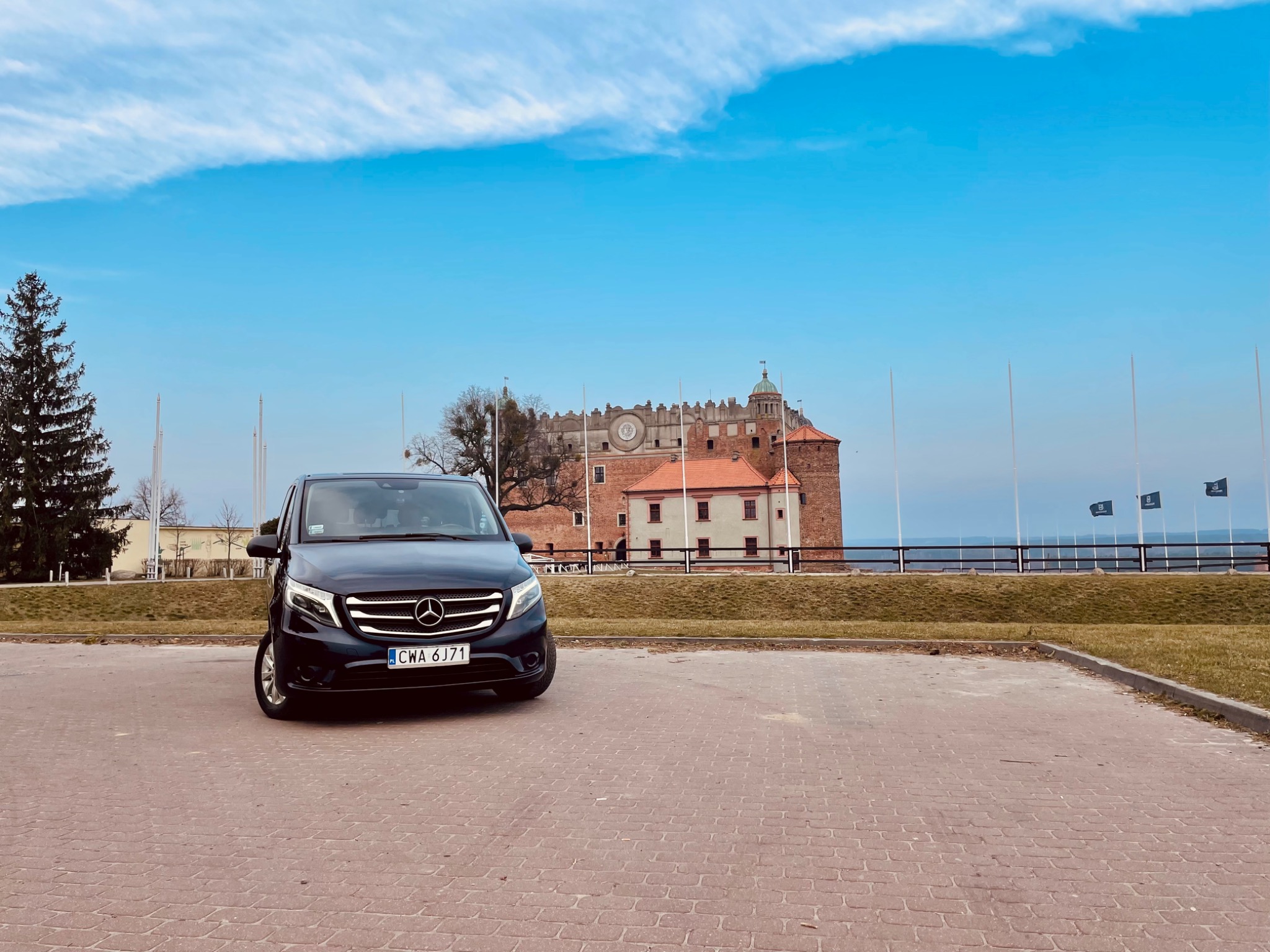 Czarny van Mercedes z numerem rejestracyjnym CWA 6J71 zaparkowany na brukowanym placu z widokiem na zamek w tle, błękitne niebo z chmurami.