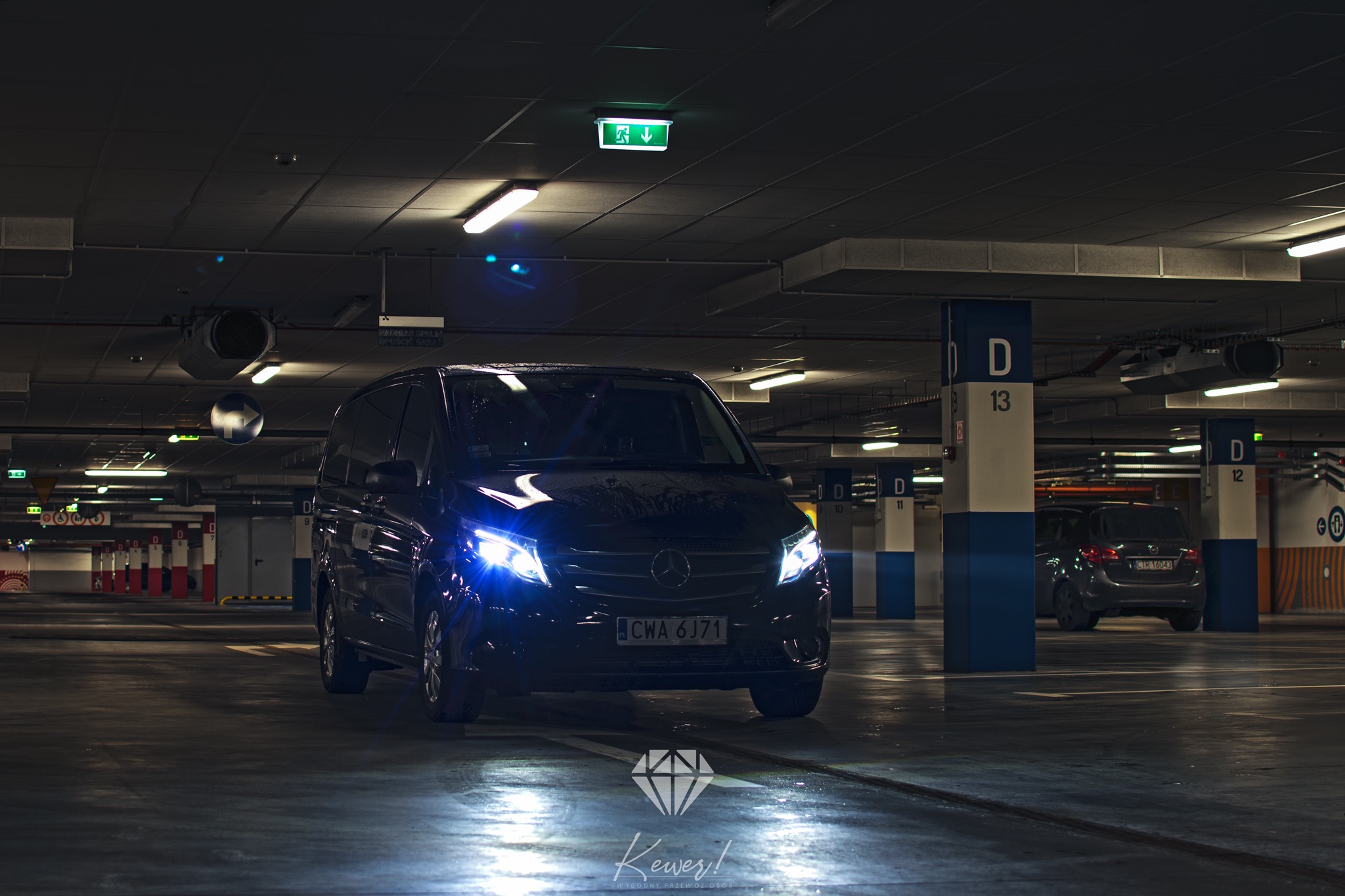 Czarny van Mercedes z włączonymi światłami na parkingu podziemnym, widoczne słupy z oznaczeniami miejsc parkingowych i znaki ewakuacyjne.