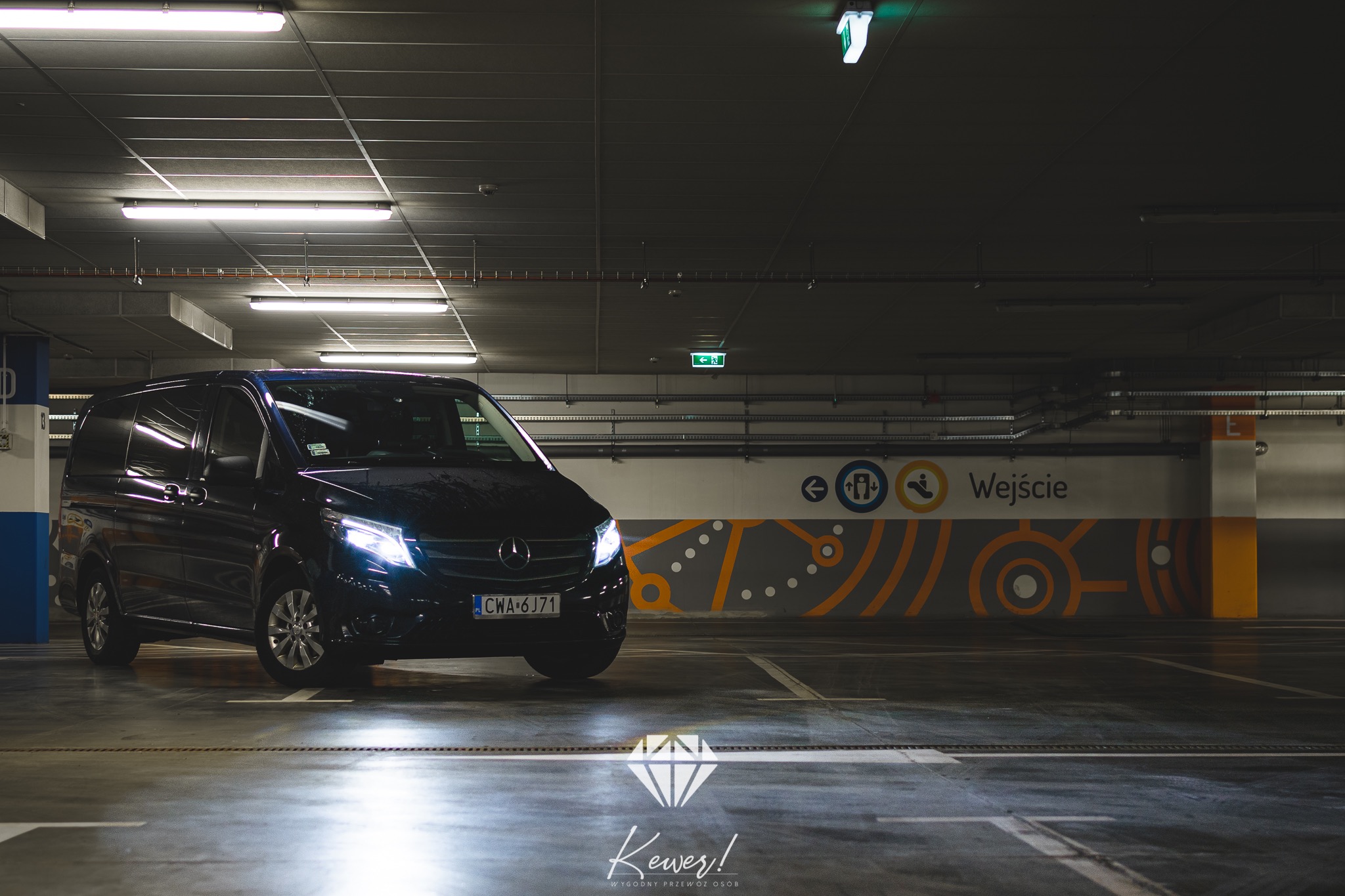 Czarny van Mercedes V-klasy z włączonymi światłami, zaparkowany w garażu podziemnym, widoczny znak 'Wejście' na ścianie.