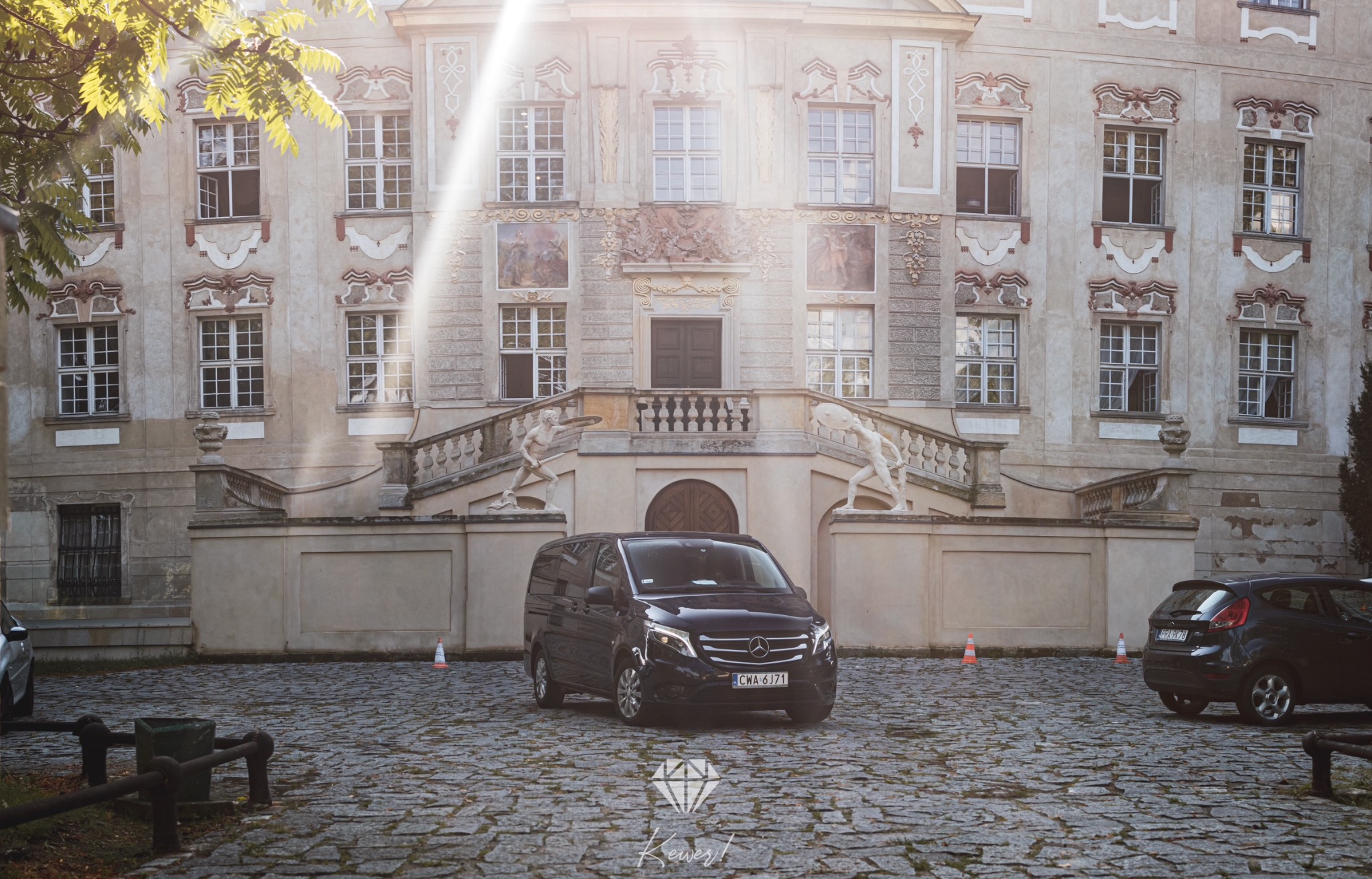 Ciemny van Mercedes-Benz zaparkowany na brukowanym dziedzińcu przed ozdobnym, jasnym budynkiem z rzeźbami i schodami.