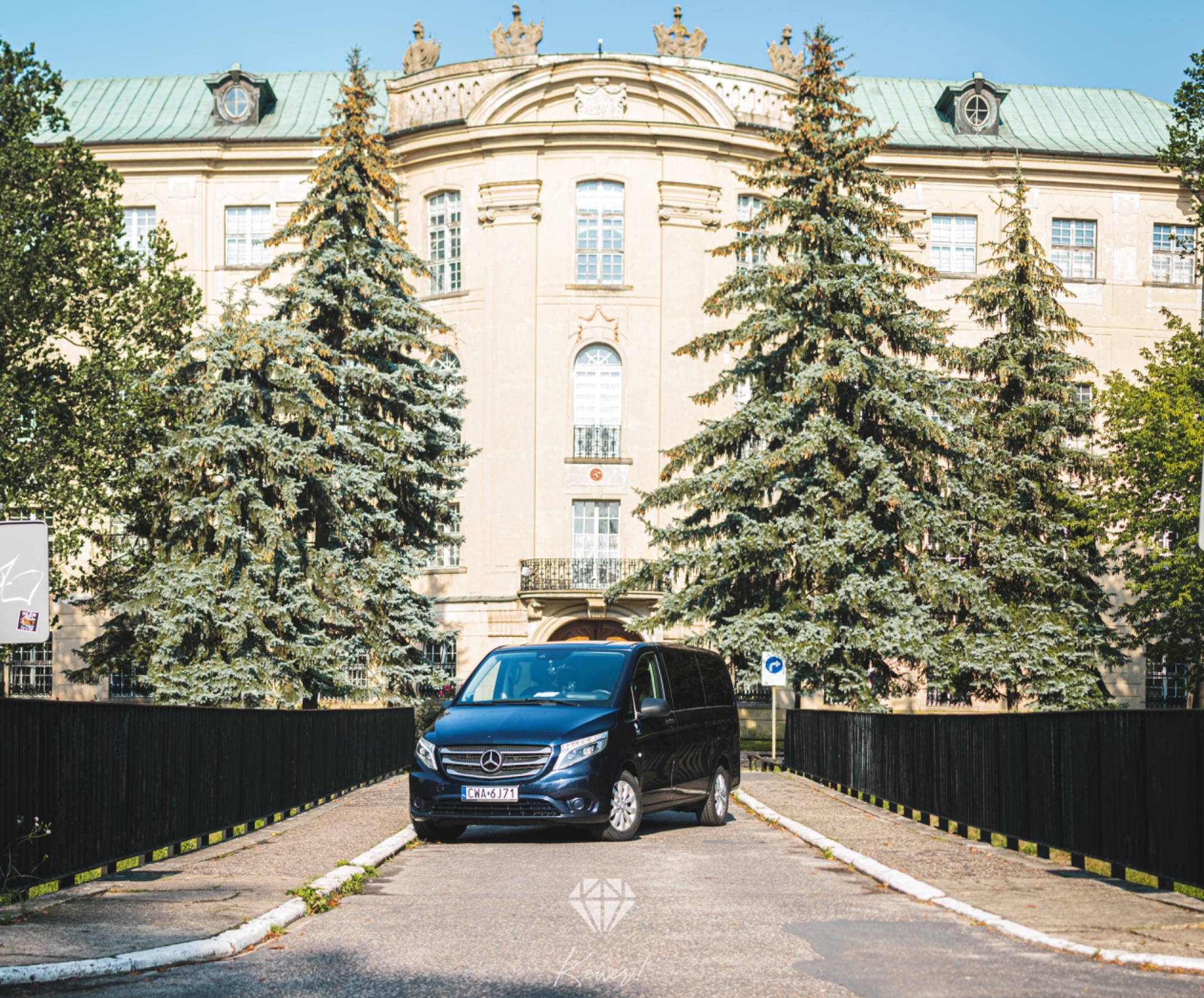 Elegancki, ciemny van Mercedes-Benz z numerami rejestracyjnymi CWA 6J71, zaparkowany na moście w Wąbrzeźnie, z klasycystycznym budynkiem w tle i wysokimi świerkami po bokach.