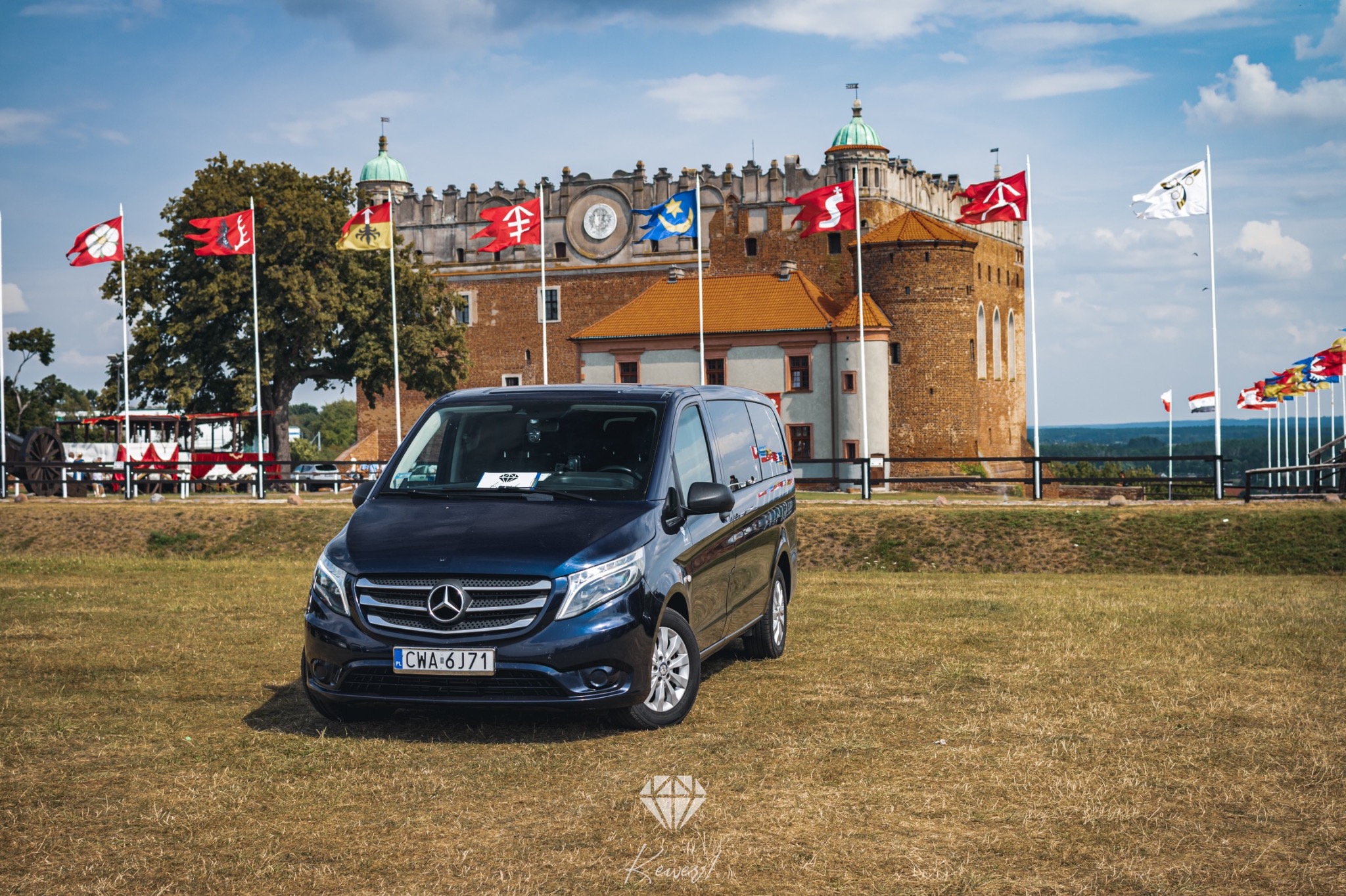 Granatowy Mercedes Vito na trawiastym placu przed zamkiem w Dobczycach, otoczonym flagami.