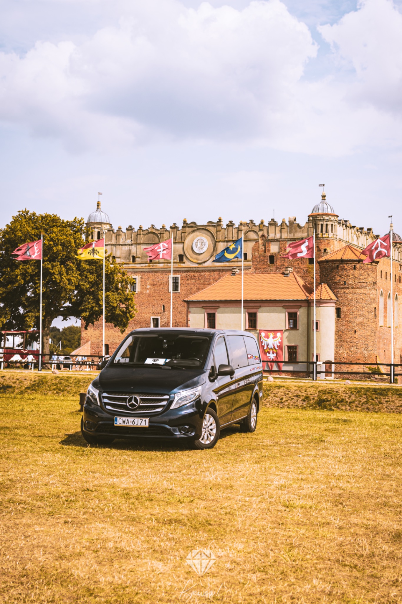 Czarny Mercedes Vito zaparkowany na trawniku przed ceglanym zamkiem z flagami, w tle błękitne niebo z chmurami.
