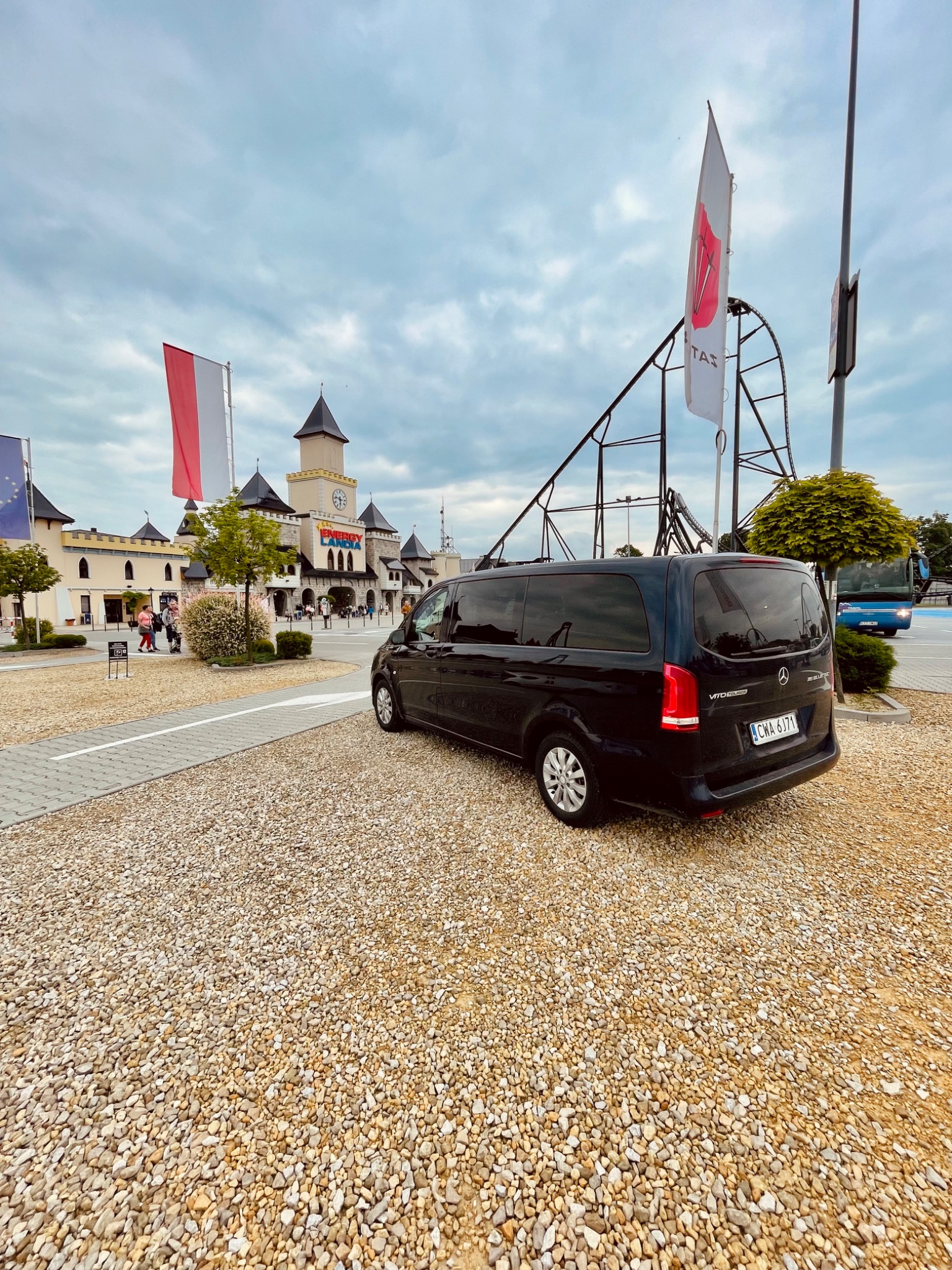 Czarny van Mercedes Vito Tourer zaparkowany na żwirowym parkingu przed parkiem rozrywki Energylandia, widoczne flagi Polski i Energylandii, w tle kolejka górska.