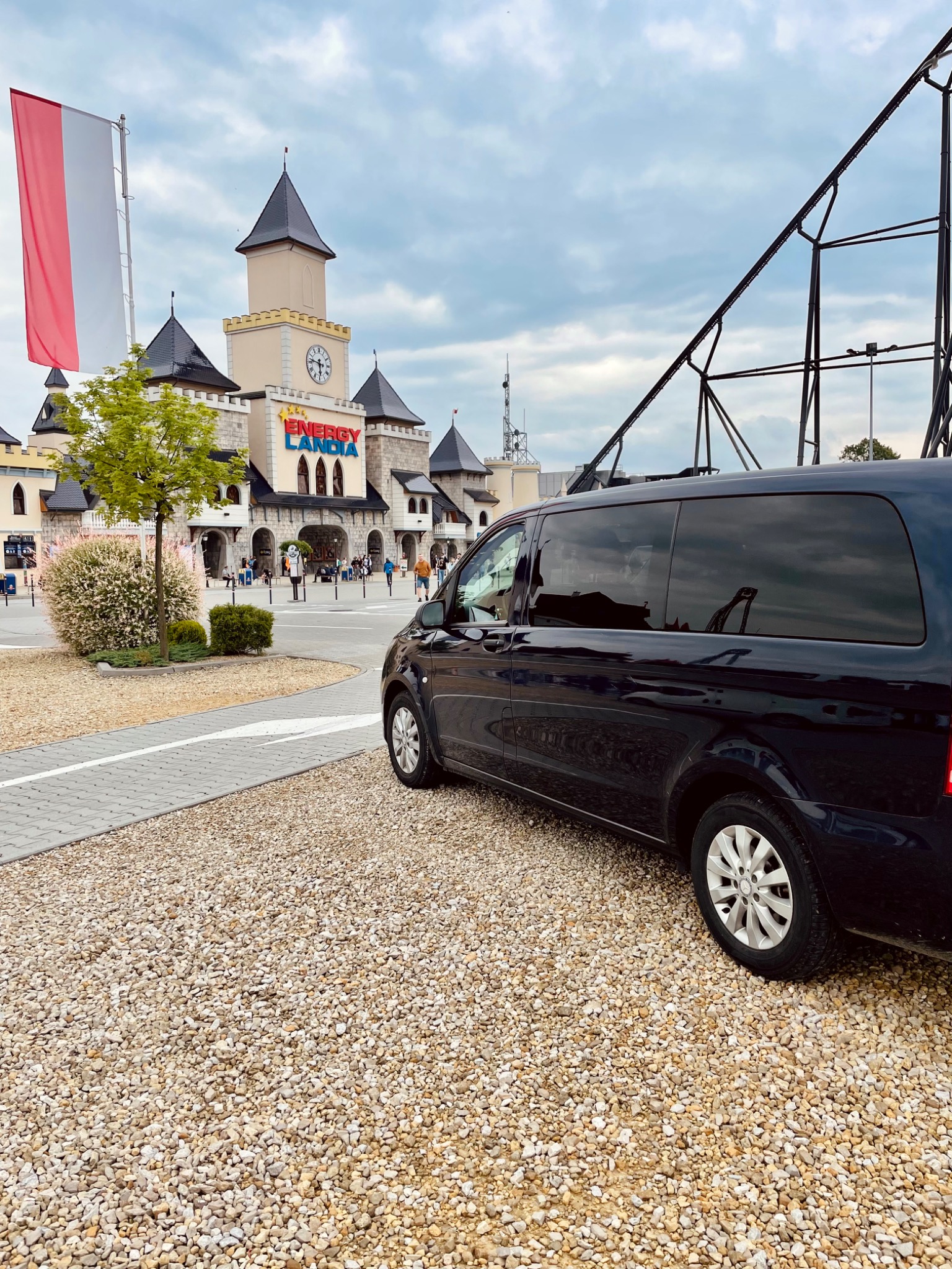 Czarny van na żwirowym parkingu przed parkiem rozrywki Energylandia, z widoczną flagą Polski w tle.