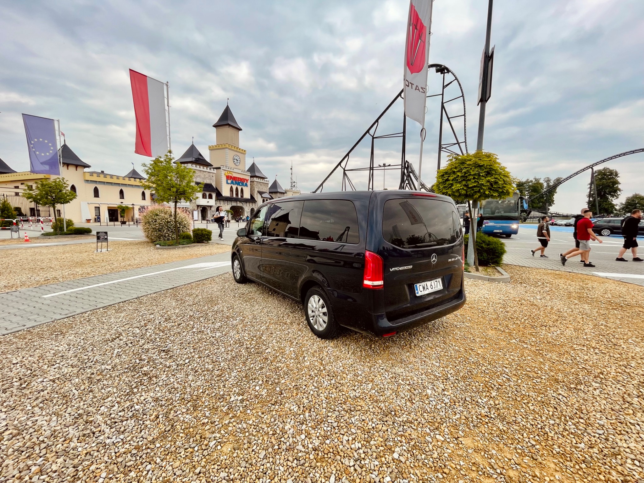Czarny van Mercedes Vito Tourer na parkingu z drobnym żwirem, w tle park rozrywki Energylandia z flagami Polski i Unii Europejskiej.