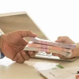 Wręczenie pliku banknotów z ręki do ręki na jasnym tle biurka z laptopem w tle, widoczne dokumenty i długopis.