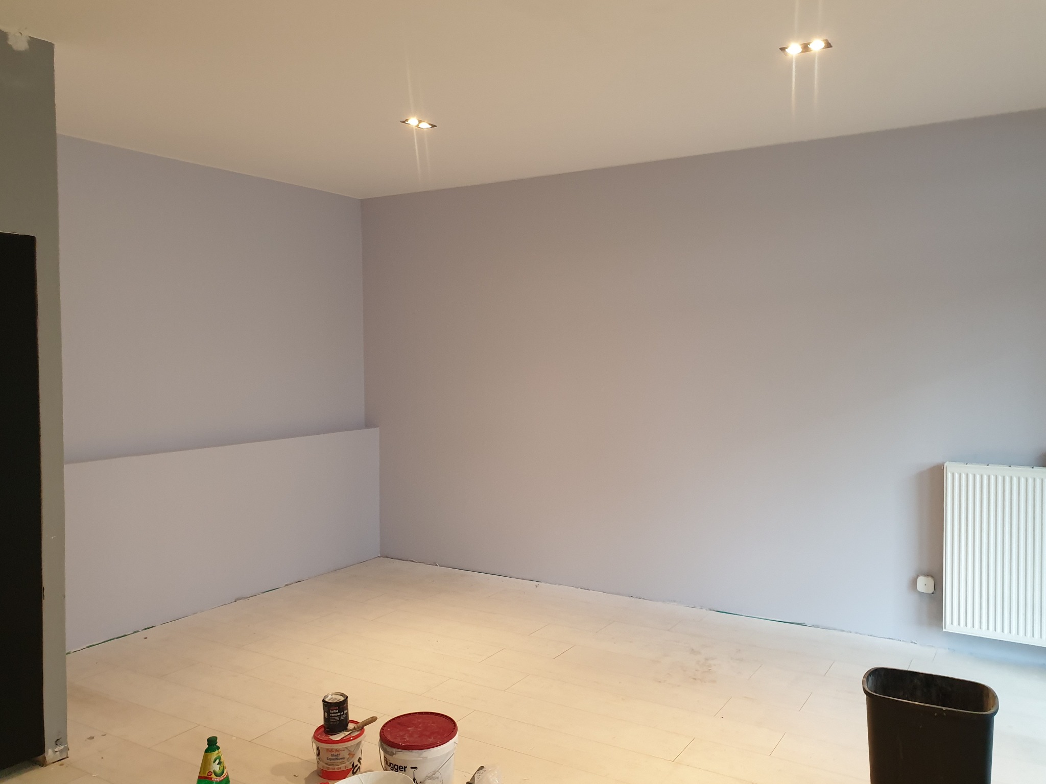 Pokój po świeżym pomalowaniu na fioletowo-szary odcień, widoczne białe podłogi, grzejnik i puszki z farbą na podłodze, przygotowane do dalszych prac.