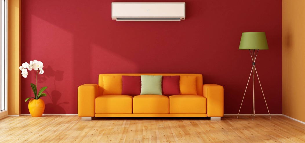 Salon z pomarańczową sofą, czerwono-pomarańczową ścianą, klimatyzacją i lampą na trójnogu, wazonie z orchideą przy oknie.