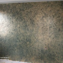 Rad-rud-system - Ściana z dekoracyjną, strukturalną fakturą w odcieniach zieleni i beżu, imitująca naturalny kamień lub marmur, z widocznymi drobnymi niedoskonałościami i śladami po gniazdkach elektrycznych.